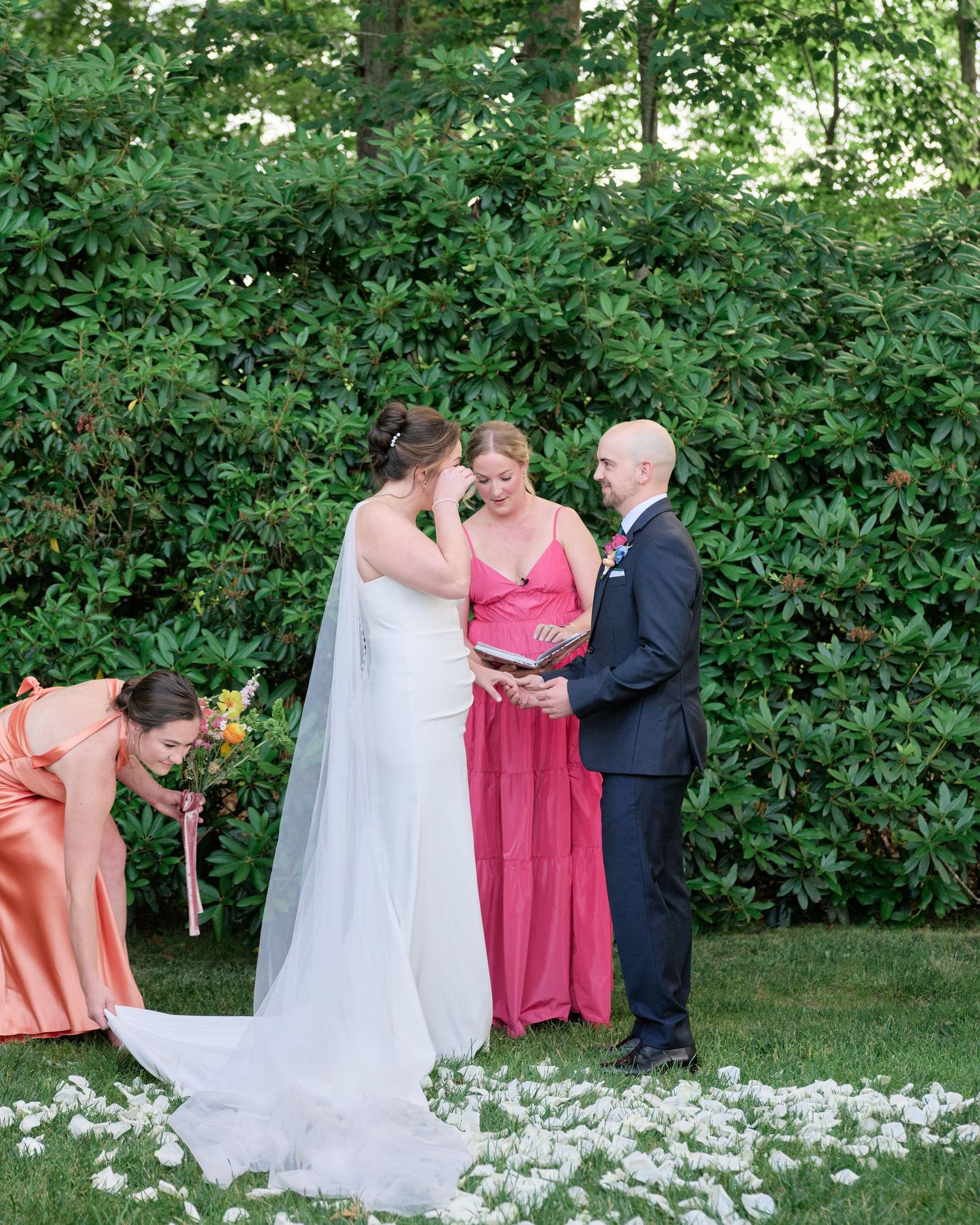 Wedding ceremony moment captured in front of green hedge with scattered flower petals.