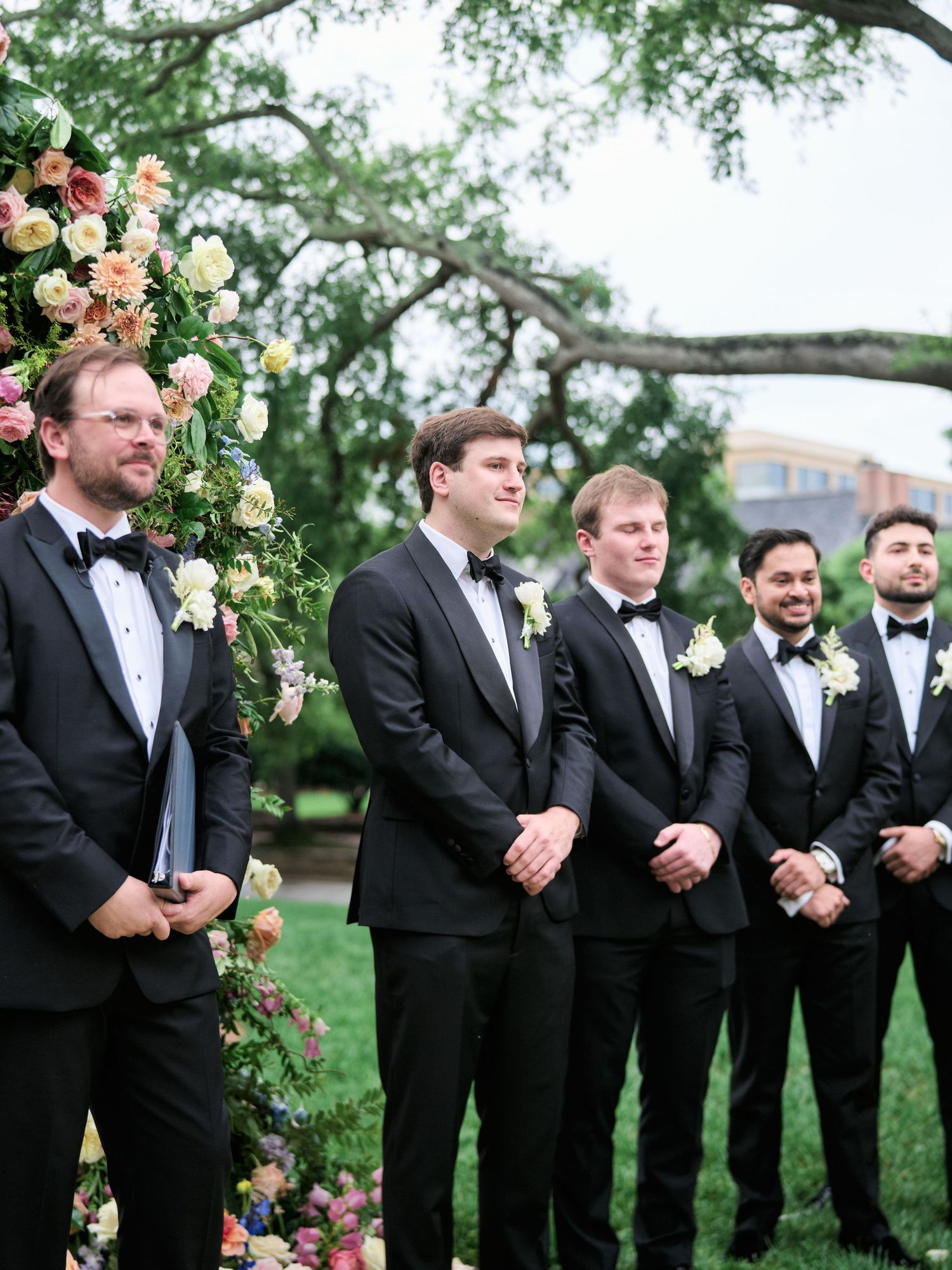 Groomsmen in black tuxedos standing under floral arch during garden wedding ceremony.
