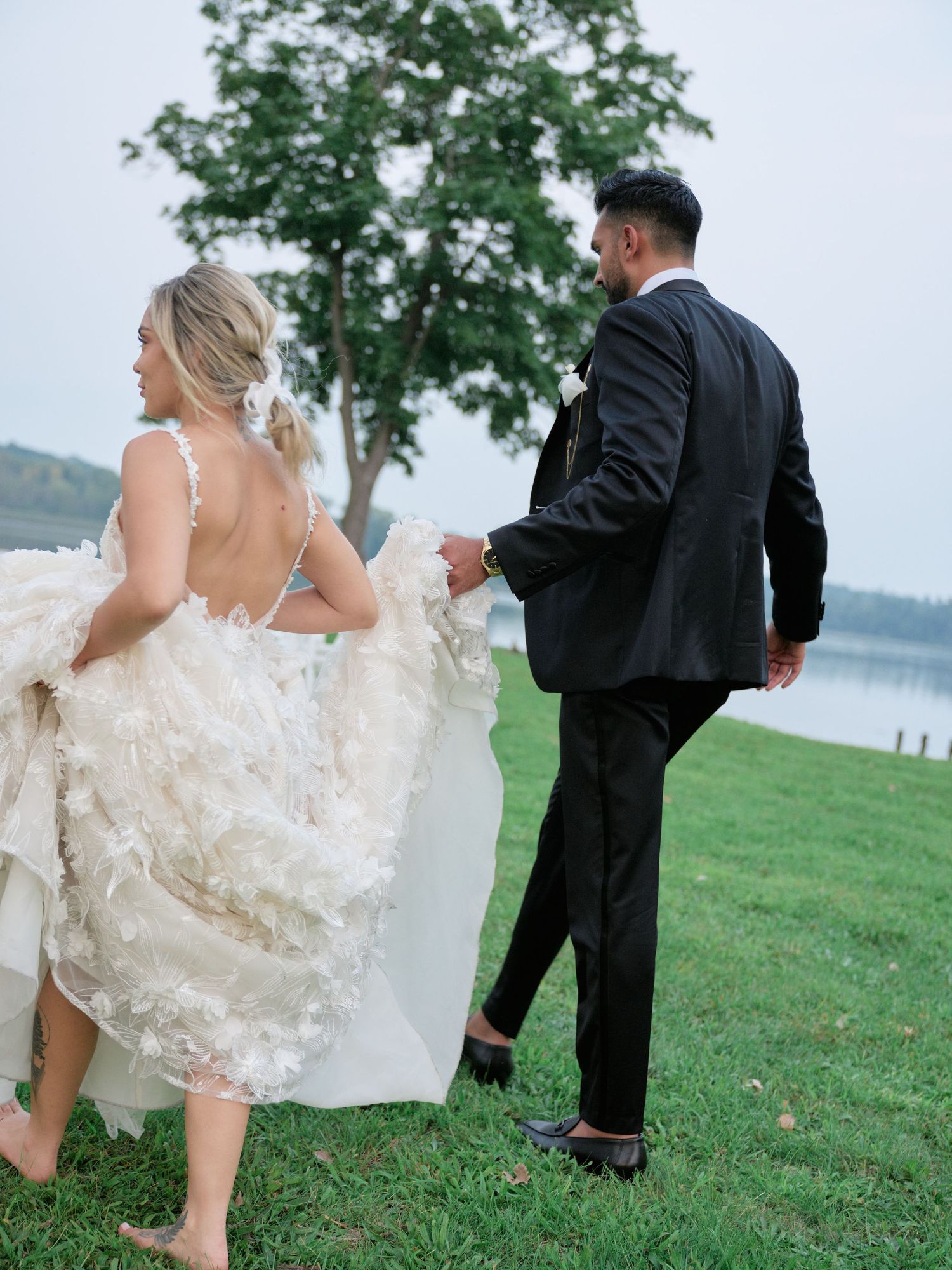 A couple in wedding attire playfully runs on a grassy hill overlooking a scenic lake on their special day.