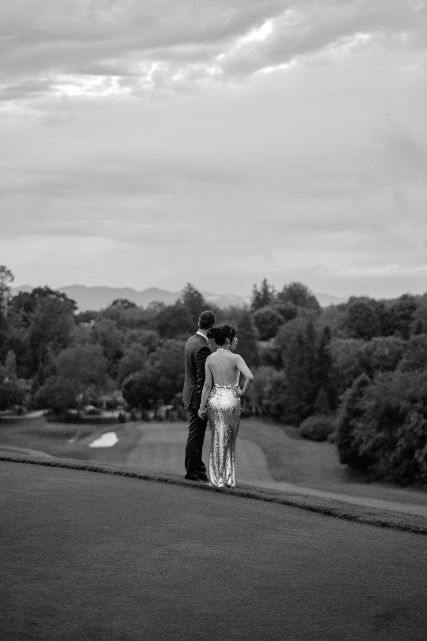 A black and white image of newlyweds standing on a hill overlooking a scenic golf course landscape.
