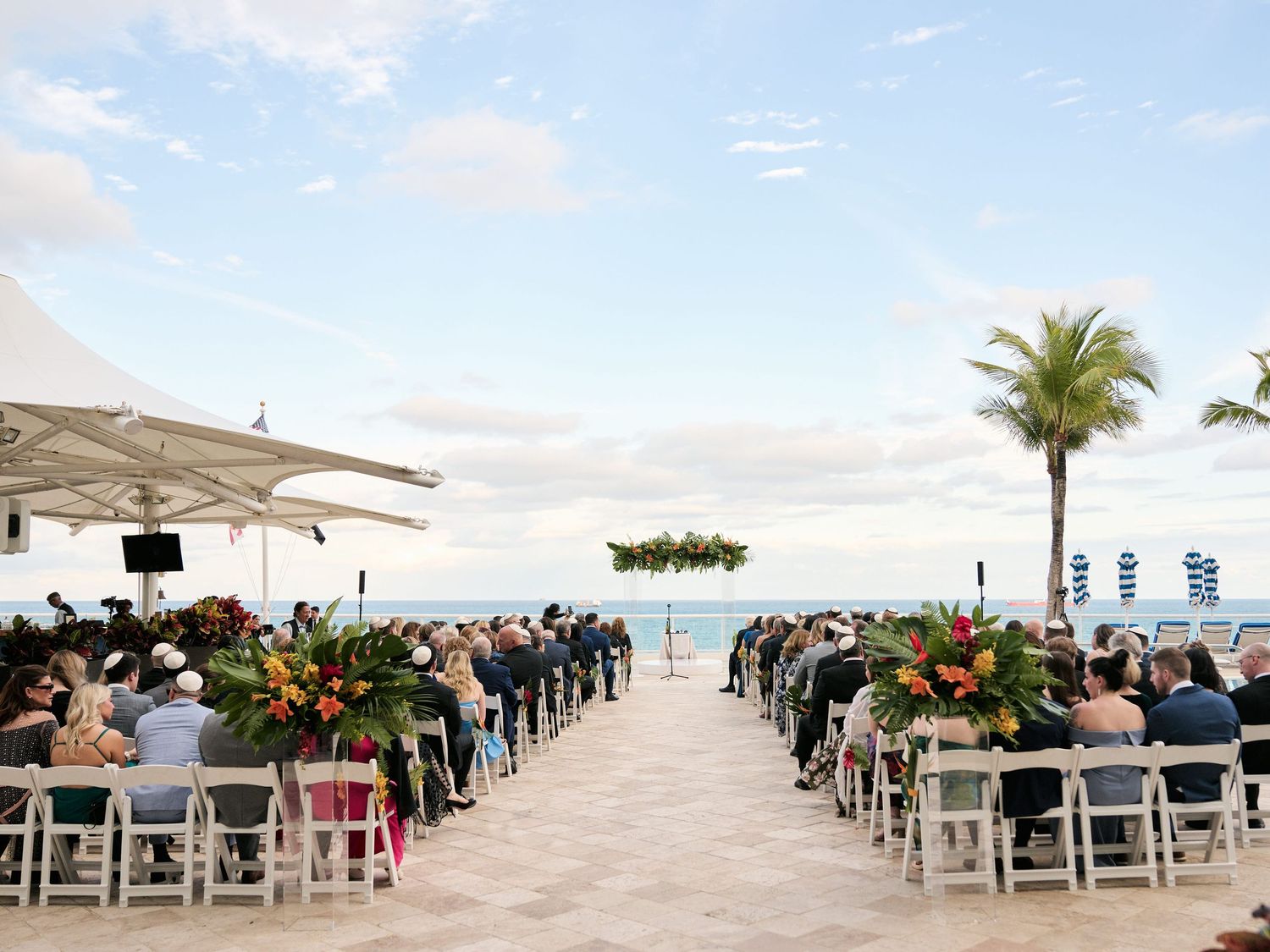 Beachfront wedding ceremony setup with white chairs and floral decorations facing the ocean.