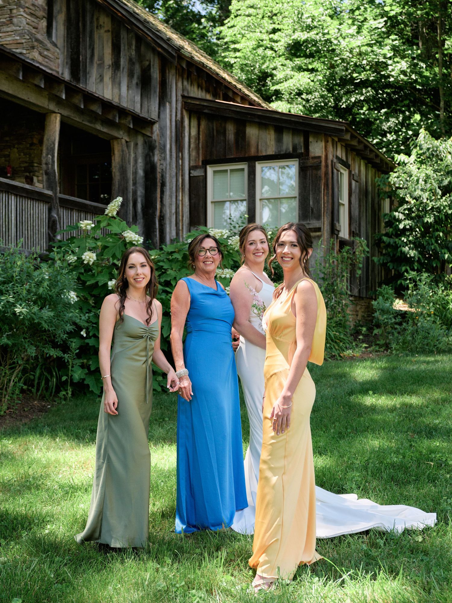 A group of bridesmaids wearing elegant floor-length dresses in various colors pose in front of a rustic cabin.