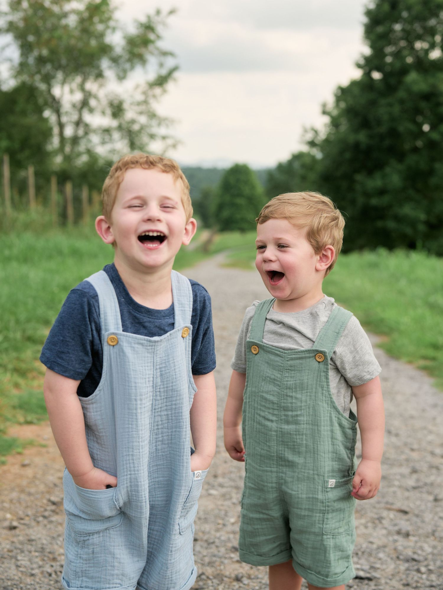 Two young children in matching overalls laugh together while standing on a gravel path in a green outdoor setting.