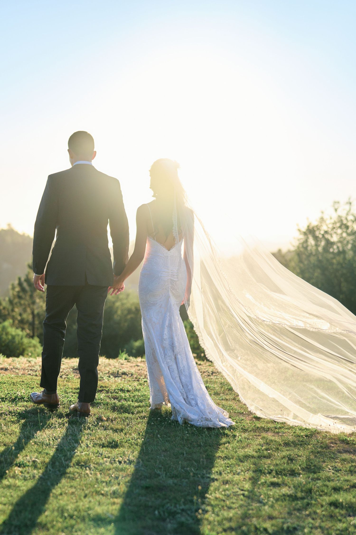 Bride and groom stand together on a hilltop during golden hour with a dramatic flowing veil.