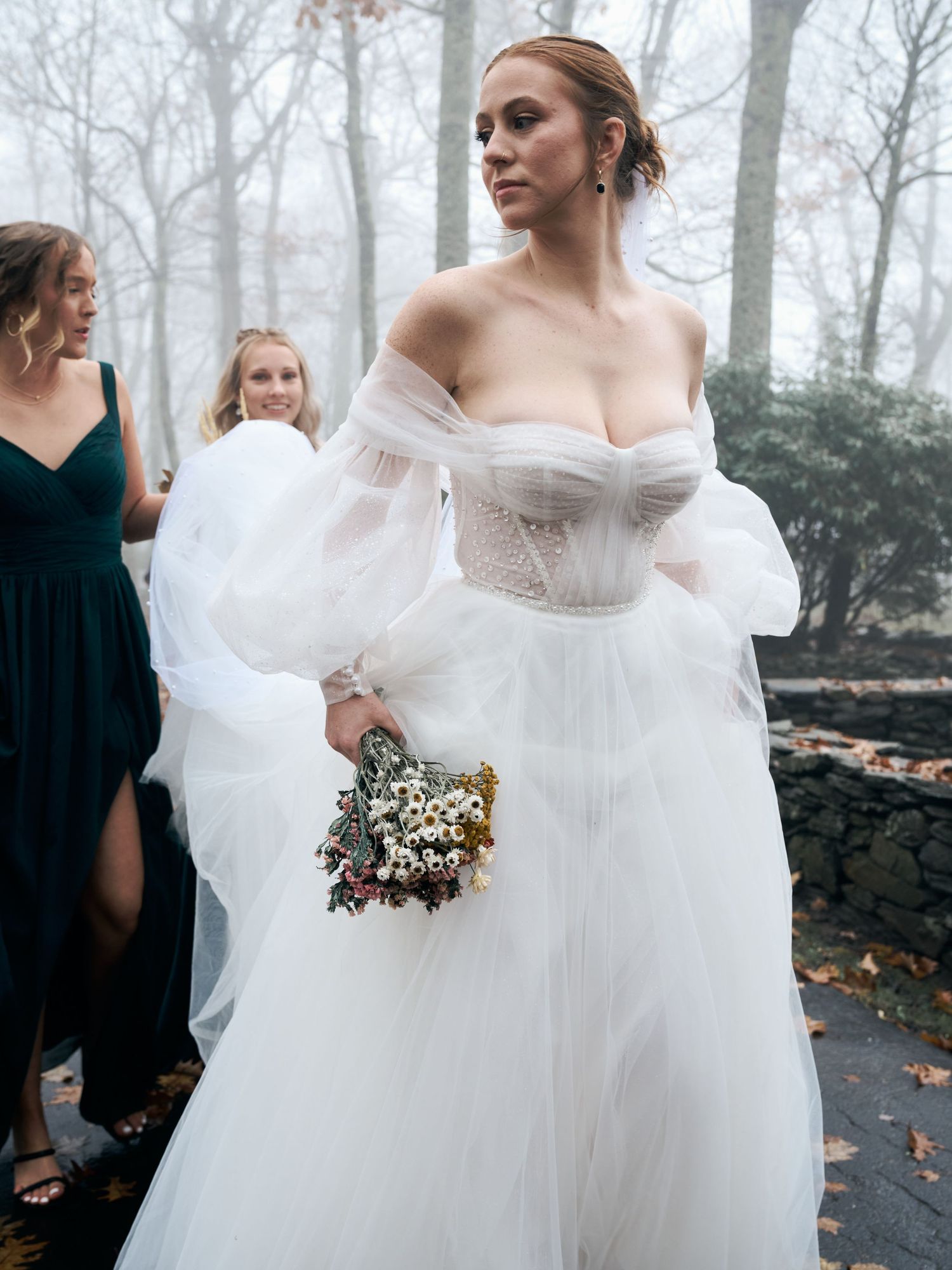 A bride in an off-shoulder white gown holds a small bouquet while standing with her wedding party.