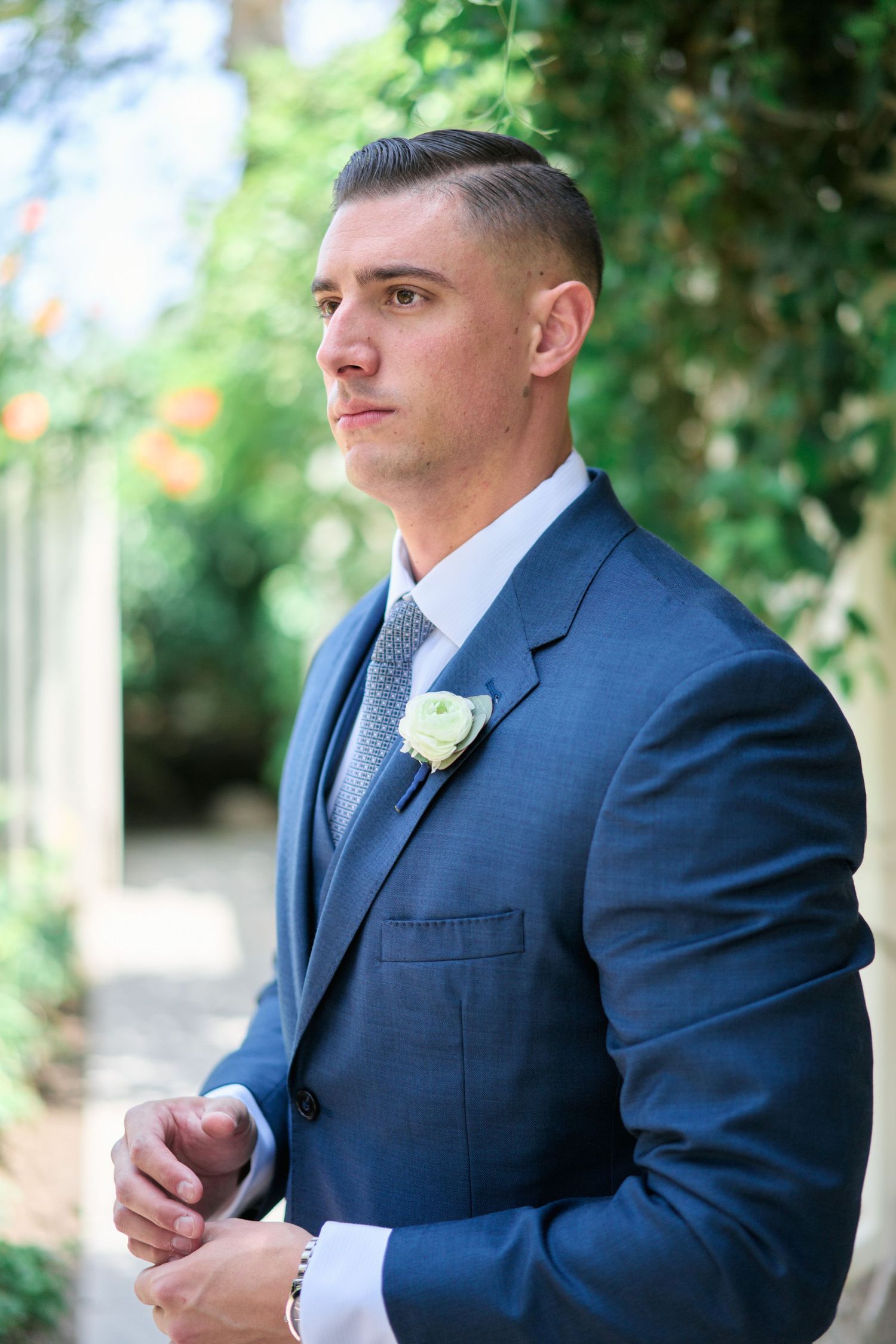 A groom in a navy blue suit with a white boutonniere poses outdoors.