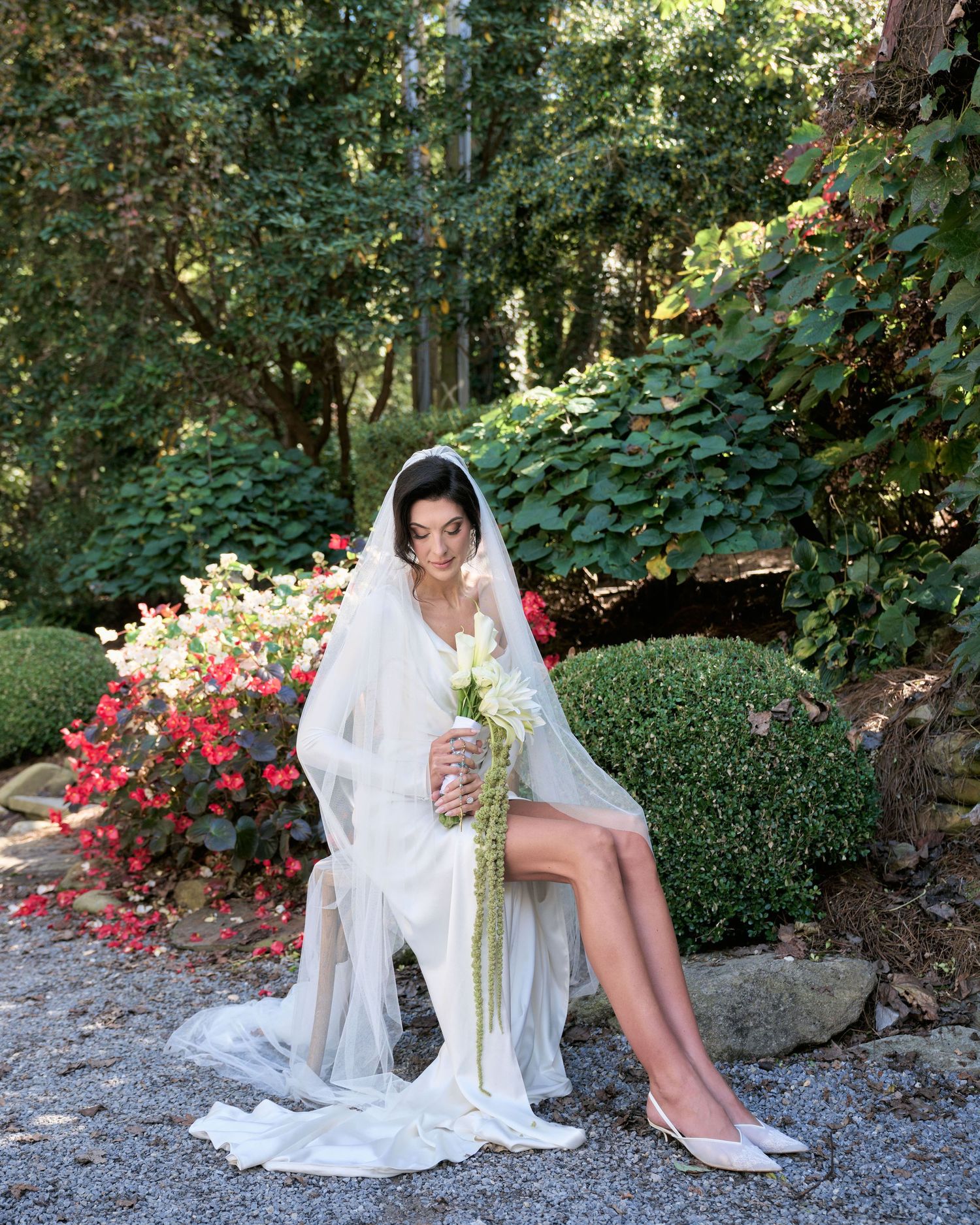 A bride in a white gown and veil sits gracefully on stone steps surrounded by lush garden foliage and pink flowers.