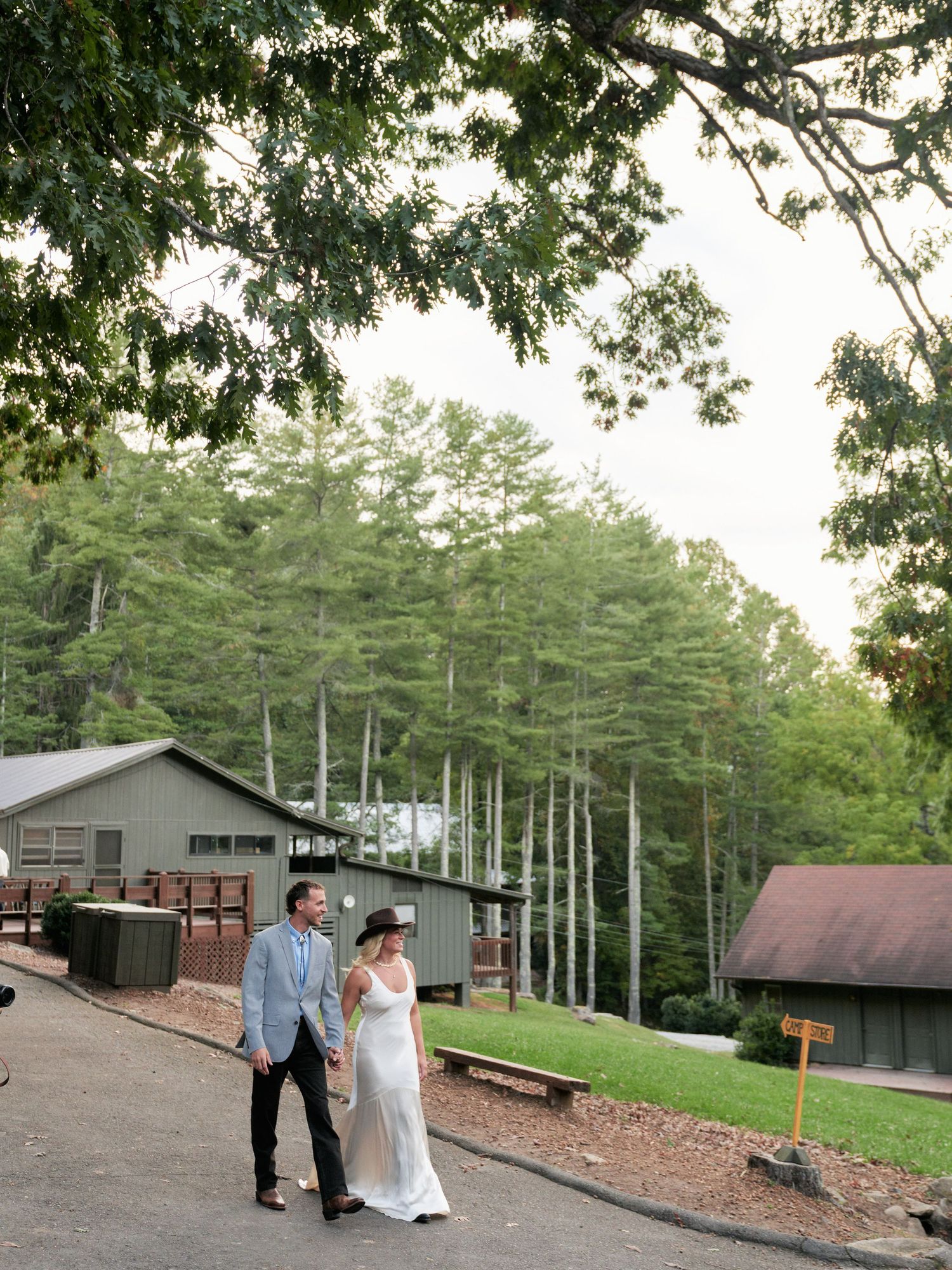 A couple shares a romantic moment on a wooded hillside with rustic buildings in the background at a summer camp venue.
