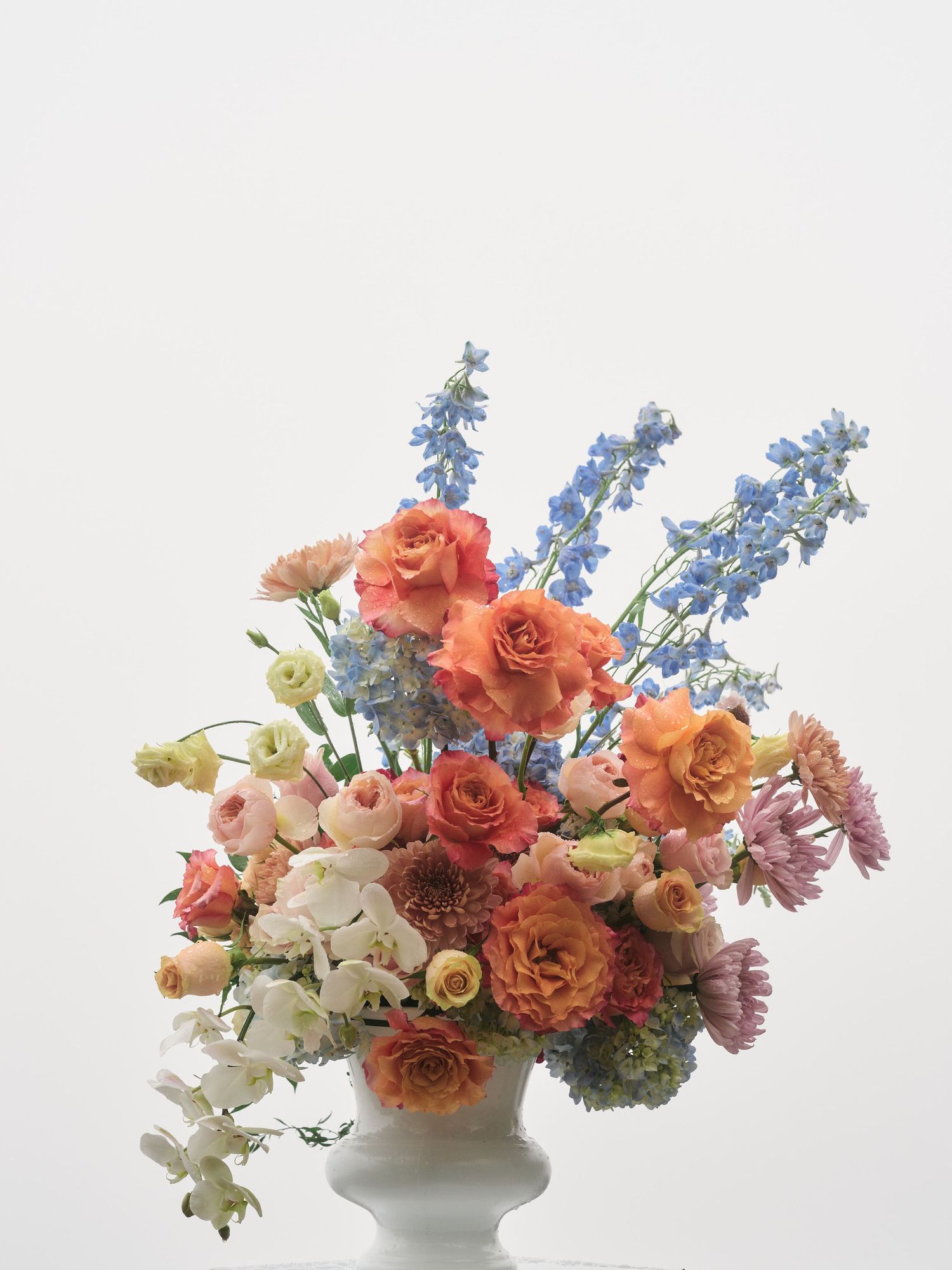 Lush floral arrangement in white urn with orange roses, blue delphiniums, pink ranunculus, and white orchids on light background.