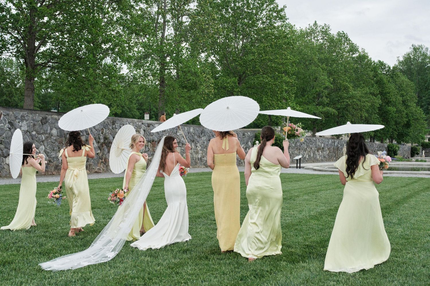 A group in light colored dresses performs a coordinated dance move in a grassy outdoor setting.