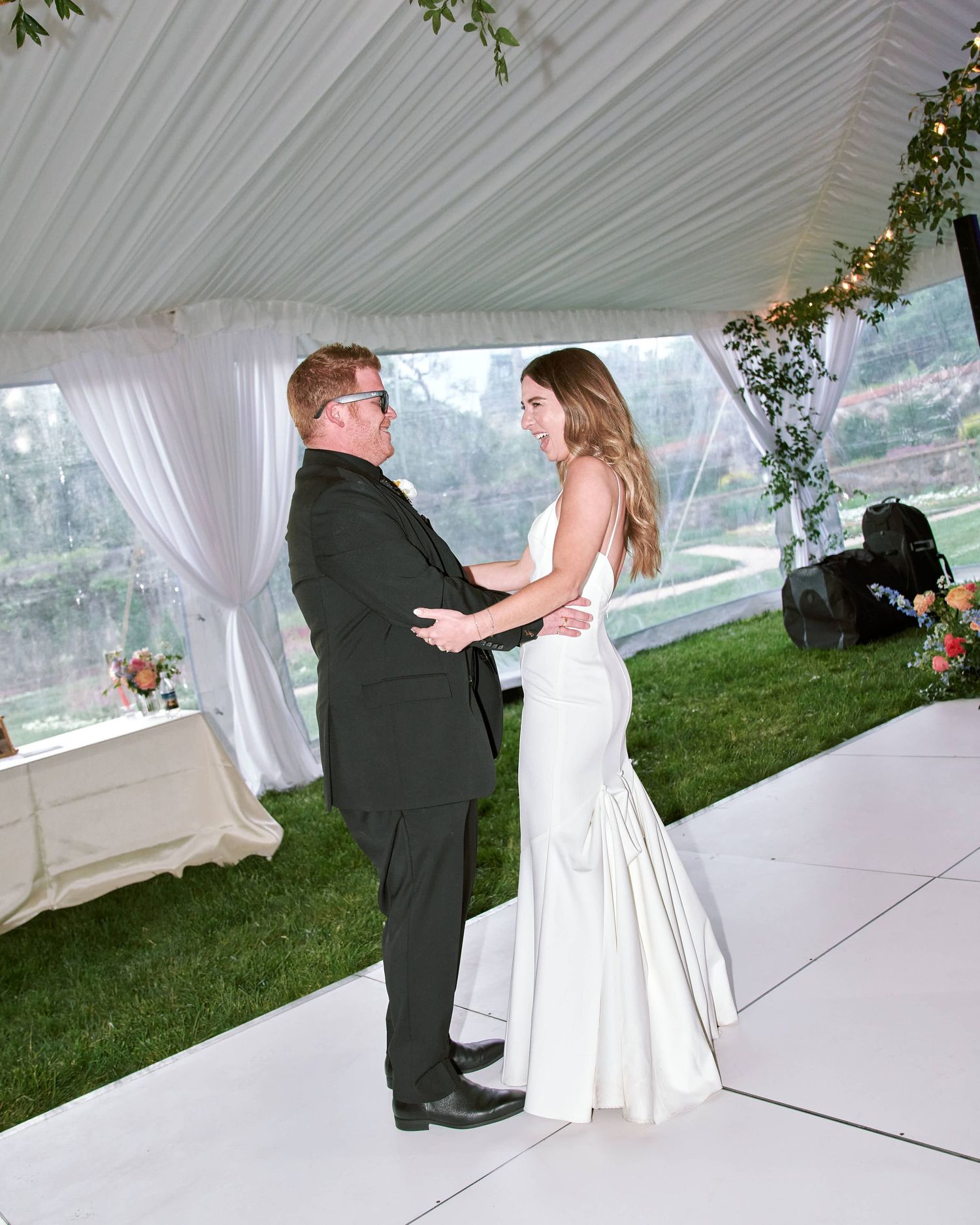 A couple performs their first dance as newlyweds under a white tent with greenery decorations.
