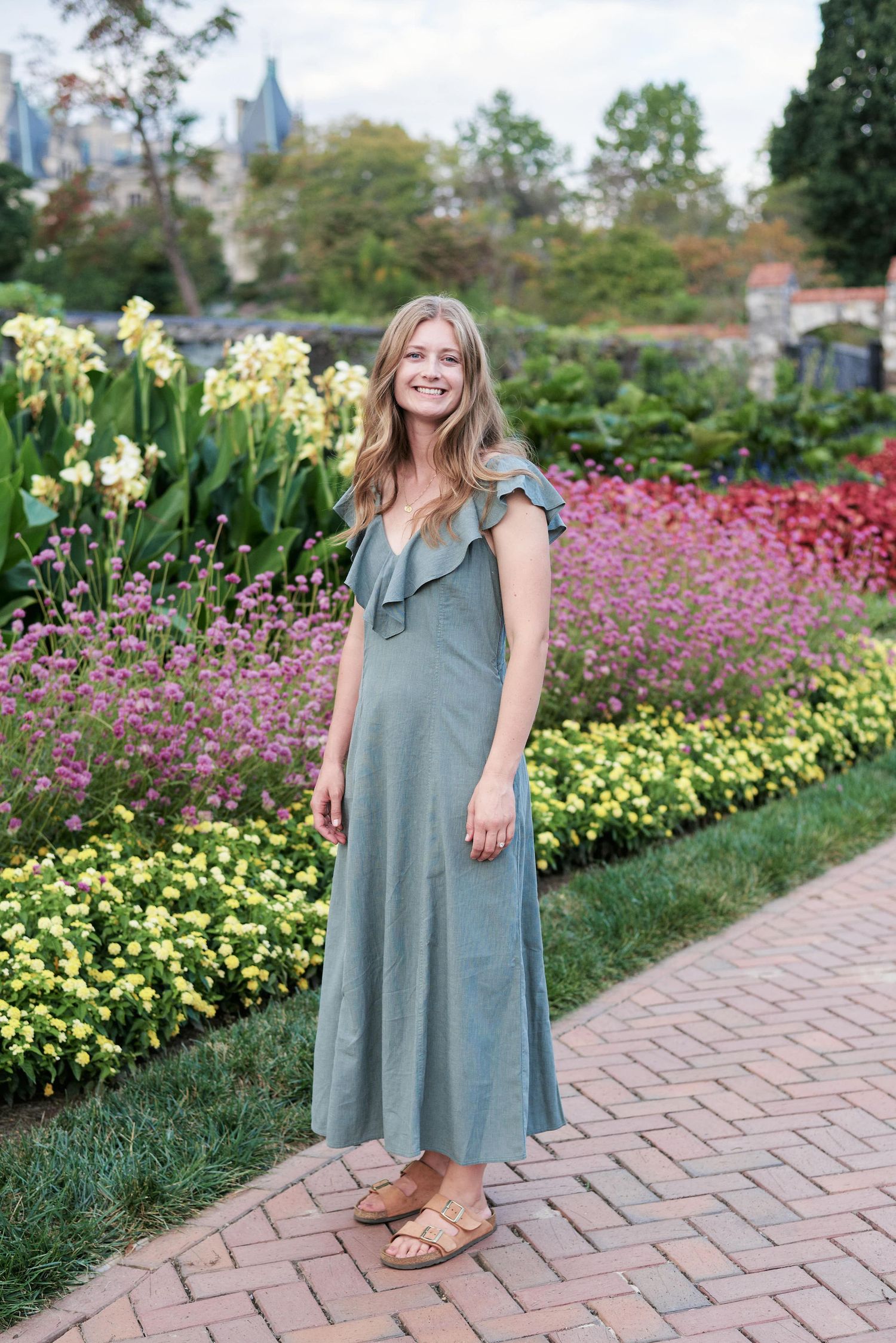 A person in a sage green dress stands on a brick path surrounded by vibrant pink and yellow flowers in a garden setting.