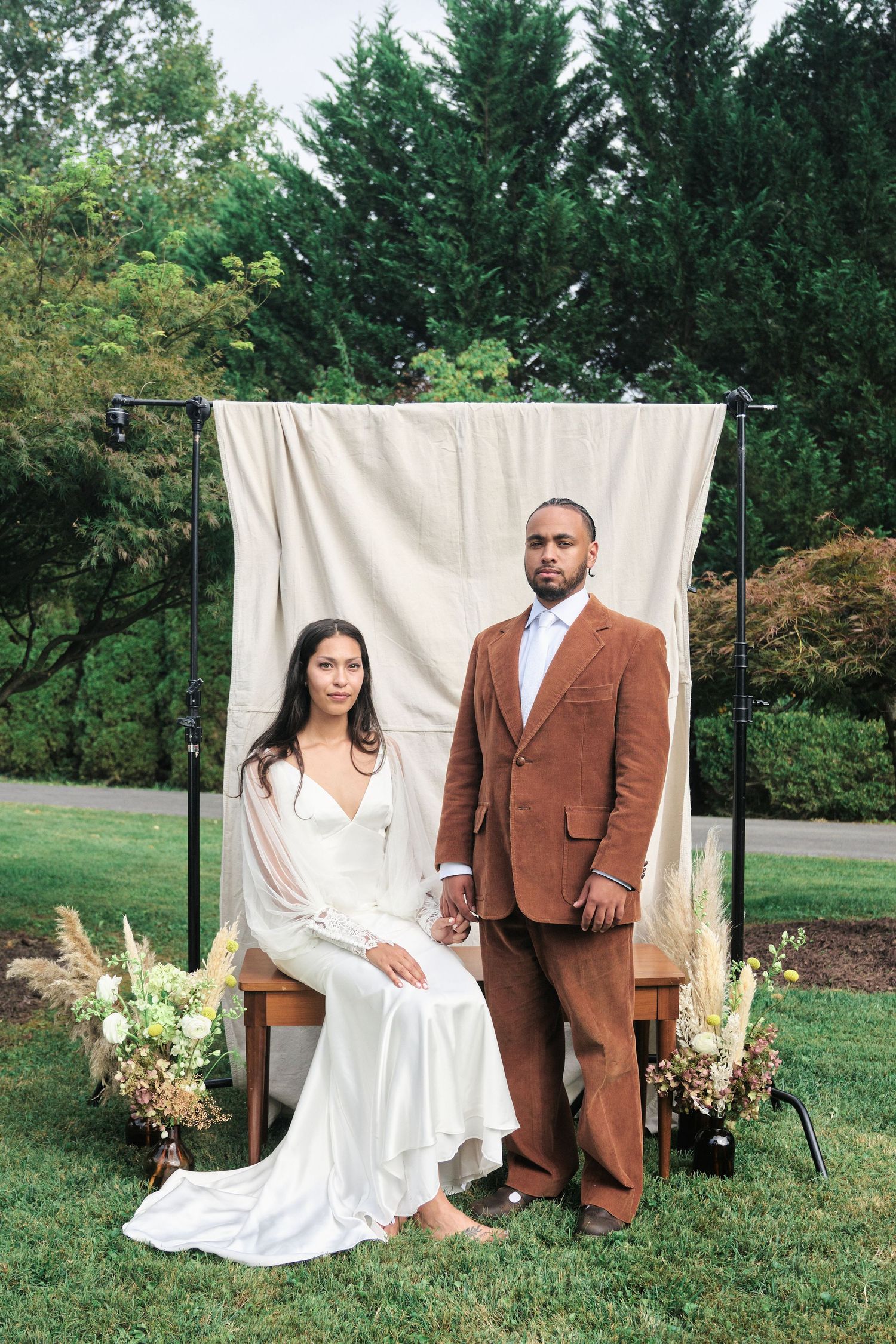 Elegant outdoor wedding portrait with couple seated against white backdrop and floral arrangements.