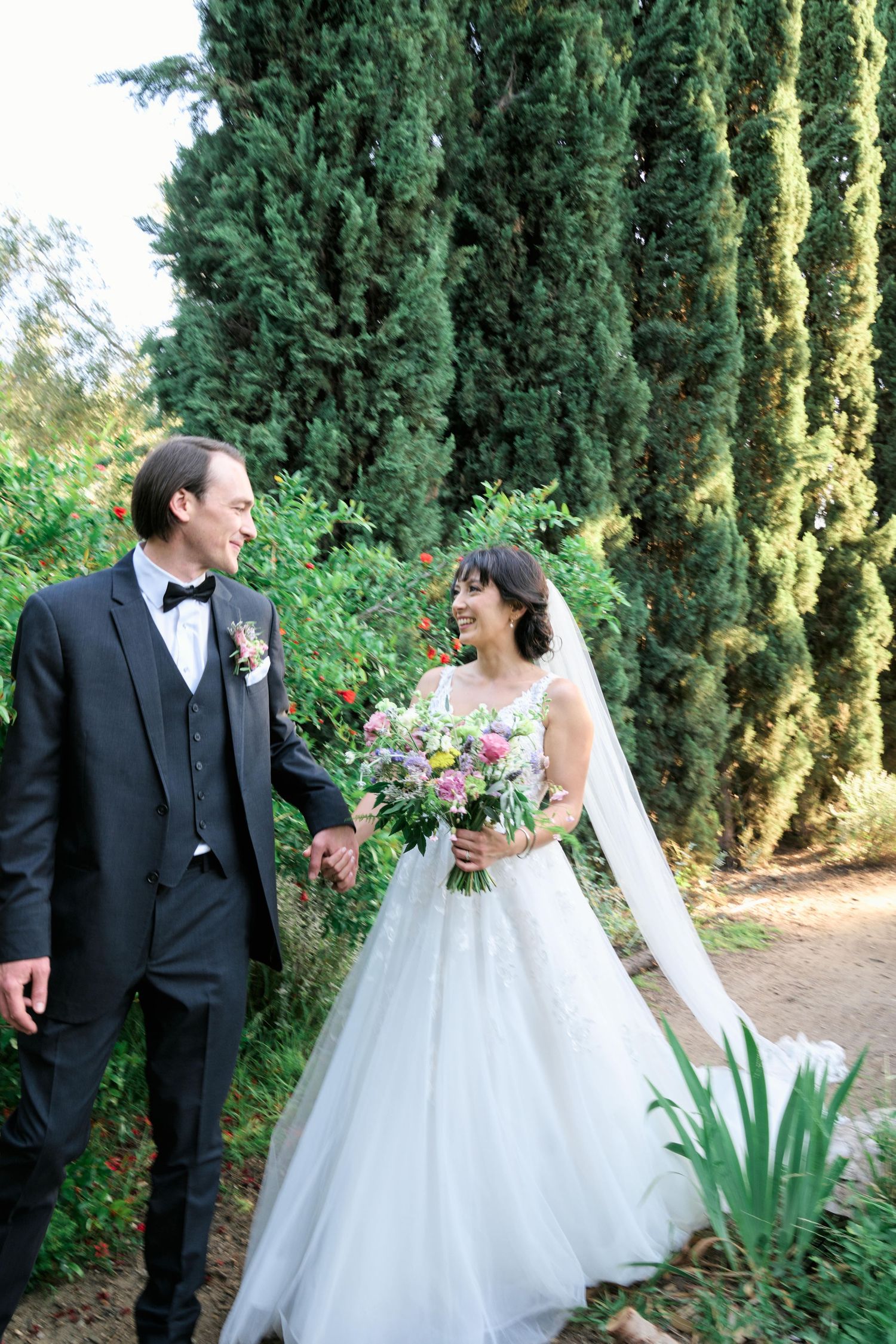 A newlywed couple walks together on a garden path lined with tall cypress trees.