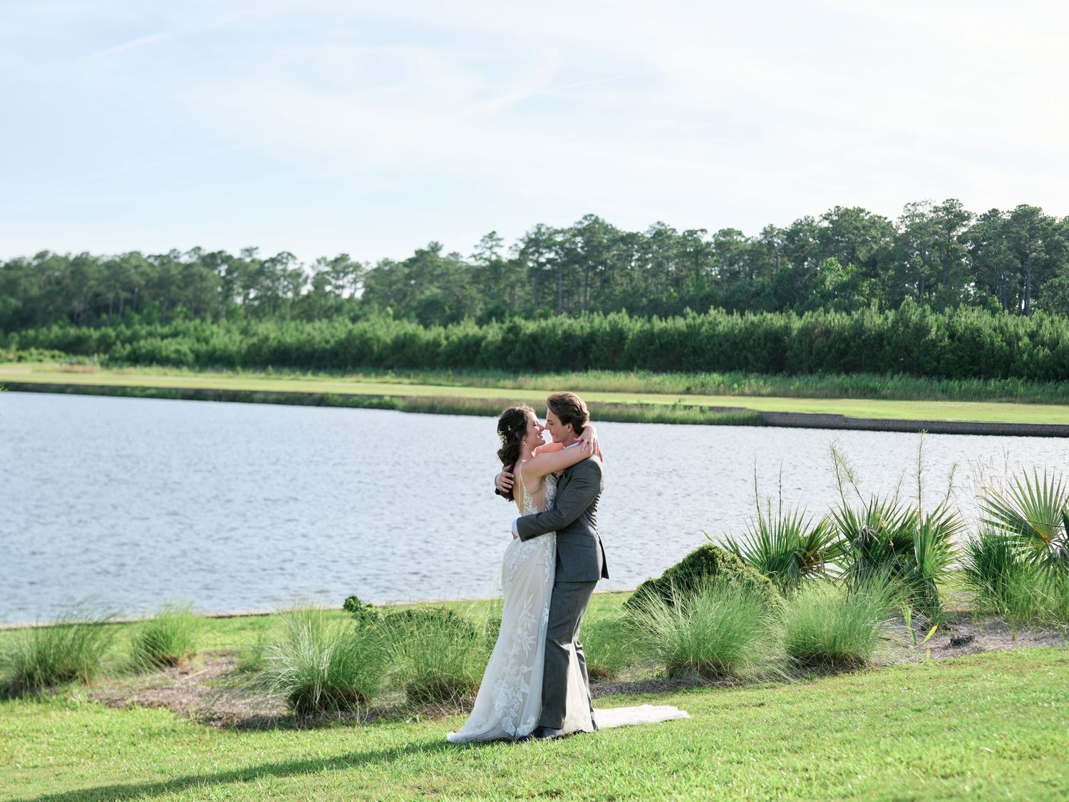 A couple embraces while standing on grassy shore beside serene lake with pine forest in the background.