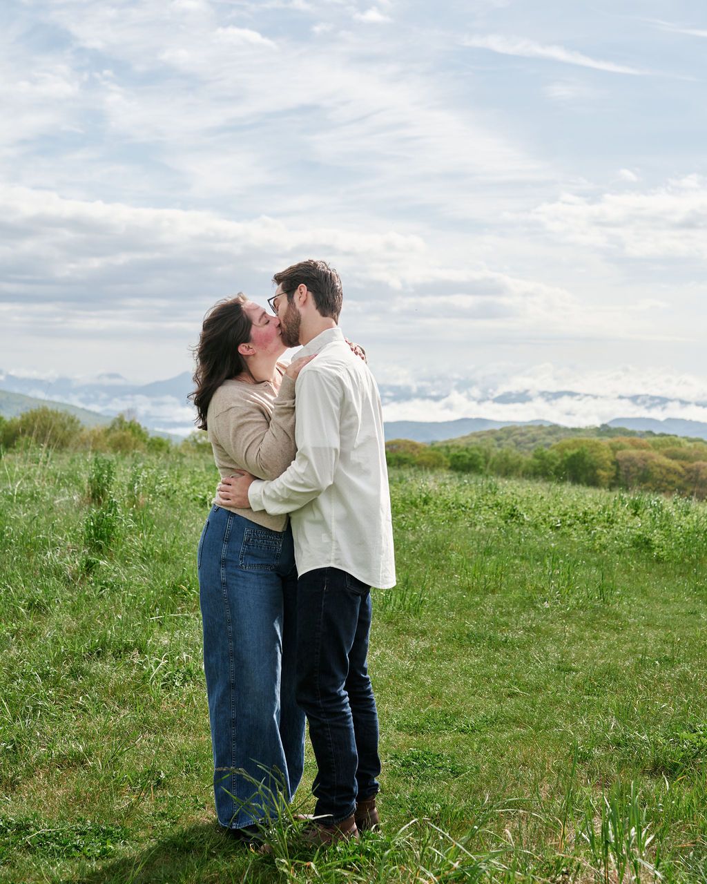 A couple embraces in a tender moment on a grassy hilltop with mountains and cloudy skies in the background.