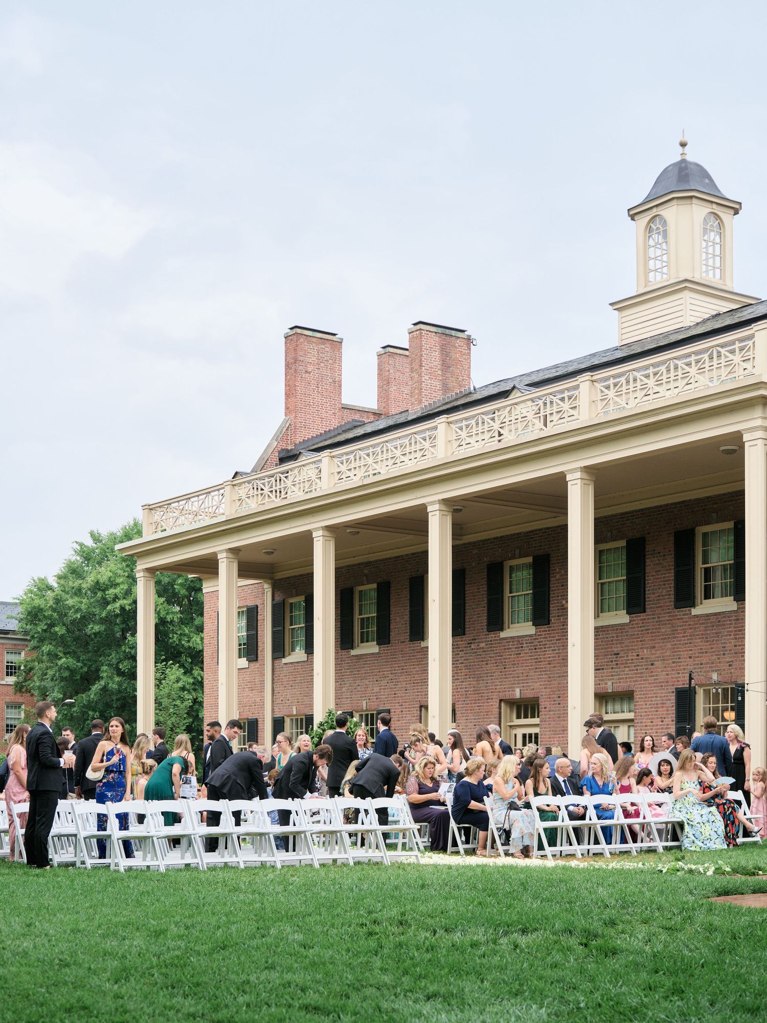 Outdoor wedding ceremony setup with white chairs on lawn in front of historic brick building with columned porch.