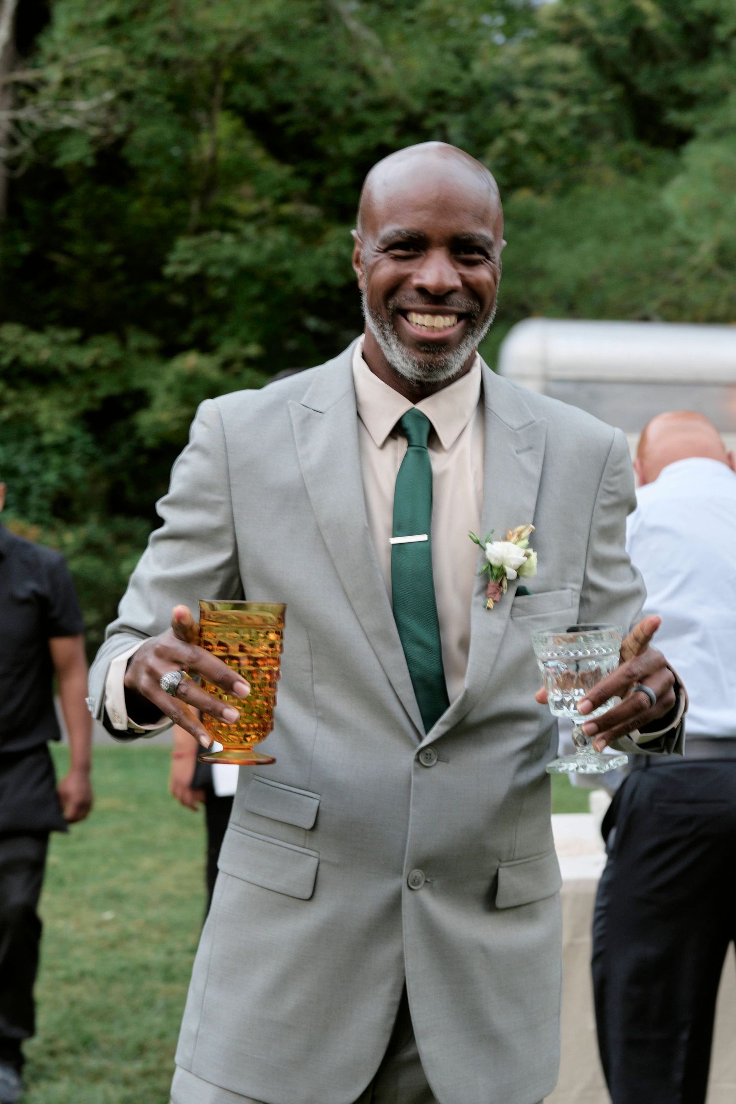 A wedding guest in a light gray suit holds two drinks while smiling at an outdoor reception.