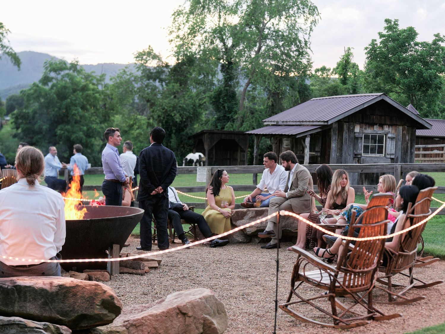 A group gathers around a fire pit at a rustic mountain lodge during dusk, with rocking chairs and cabin in background.