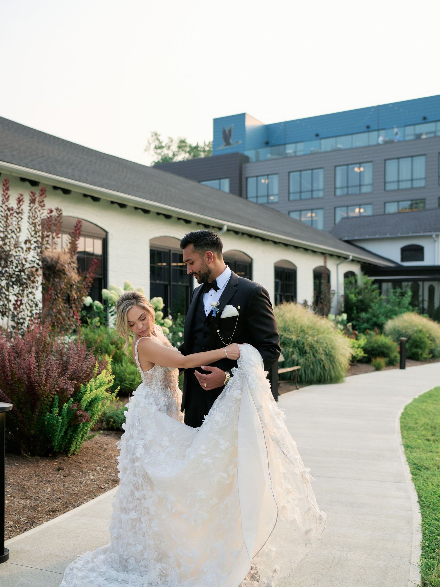 A couple in wedding attire dance and embrace on a garden path outside a building with ornamental grasses.