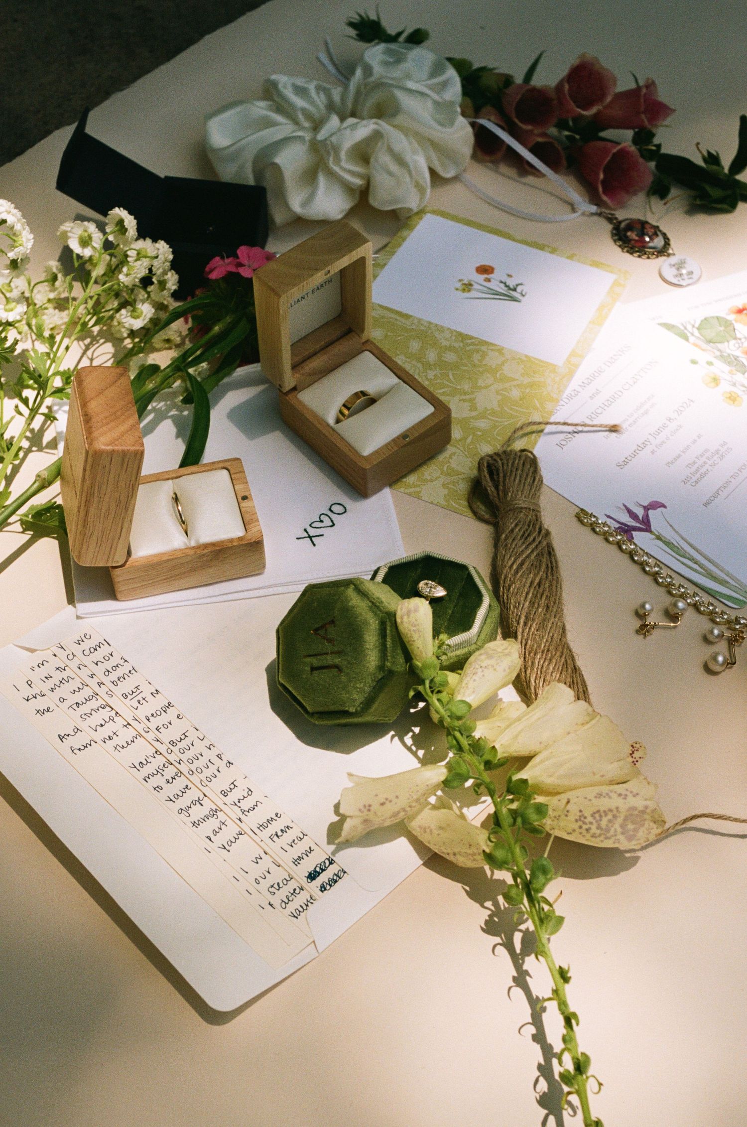 Wedding stationery and ring boxes arranged with delicate flowers and greenery in soft lighting.