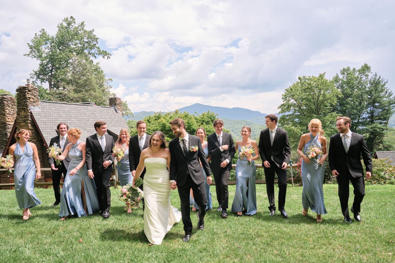 Wedding party walks outdoors on green lawn with mountain views, bride in white gown, bridesmaids in blue dresses.