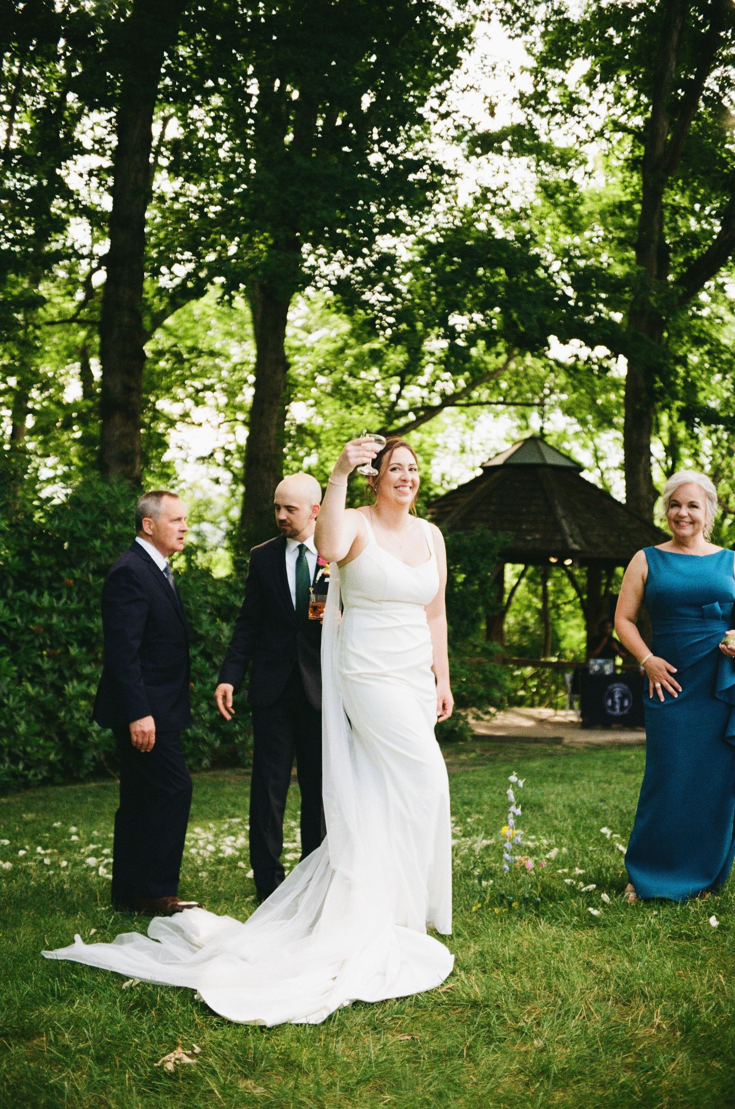 Bride in white gown laughing outdoors at garden wedding ceremony with guests and gazebo in background.