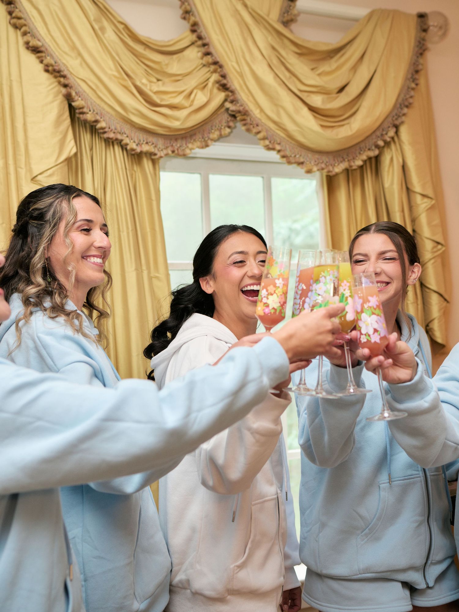 Three women in matching light blue hoodies toasting with floral champagne glasses in an elegant room with gold curtains.