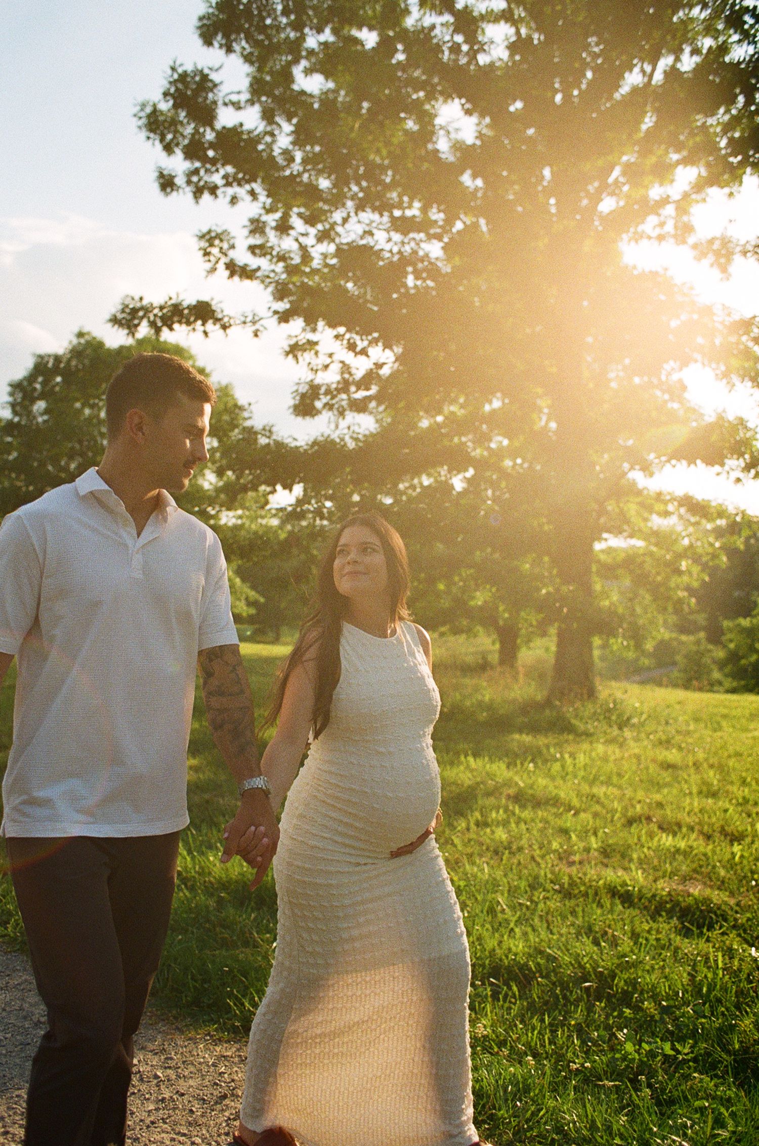 Pregnant couple holding hands walking outdoors at golden hour with sunlight glowing through trees in a lush green park.