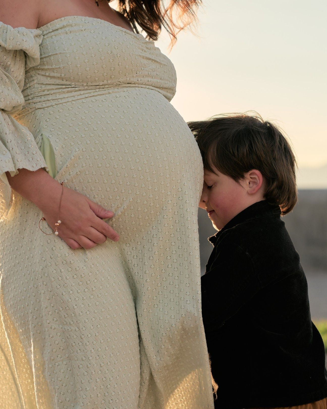 A pregnant woman in a cream dress stands as a young child gently kisses her belly during a sunset photoshoot.