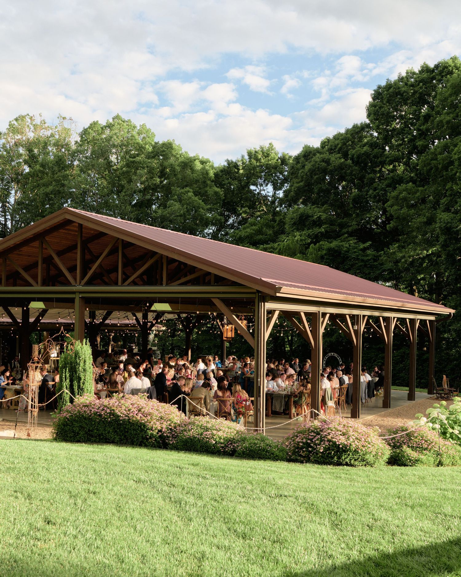 Rustic outdoor pavilion with wooden beams hosts a gathering on a grassy lawn surrounded by trees on a sunny day.