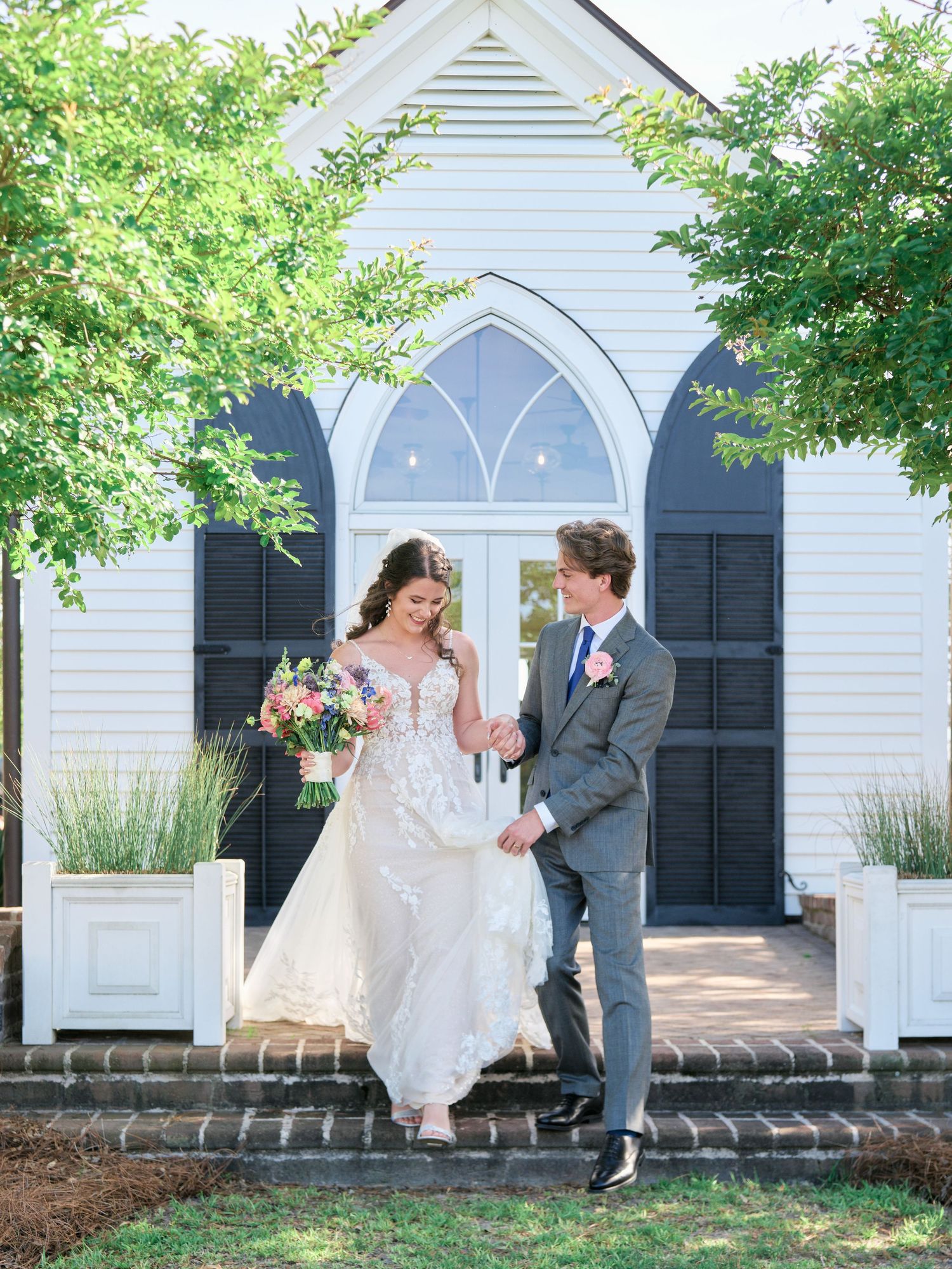 A couple exits a charming white chapel with arched Gothic windows and black shutters on a sunny summer day.