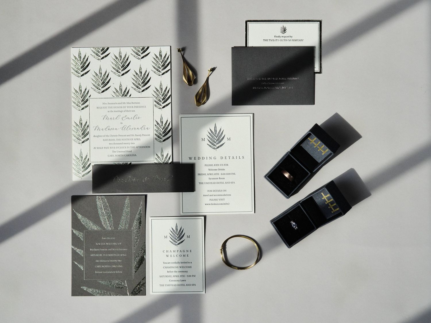 Elegant wedding stationery set with leaf motifs, black tubes, and ring displayed on white surface with dramatic shadows.