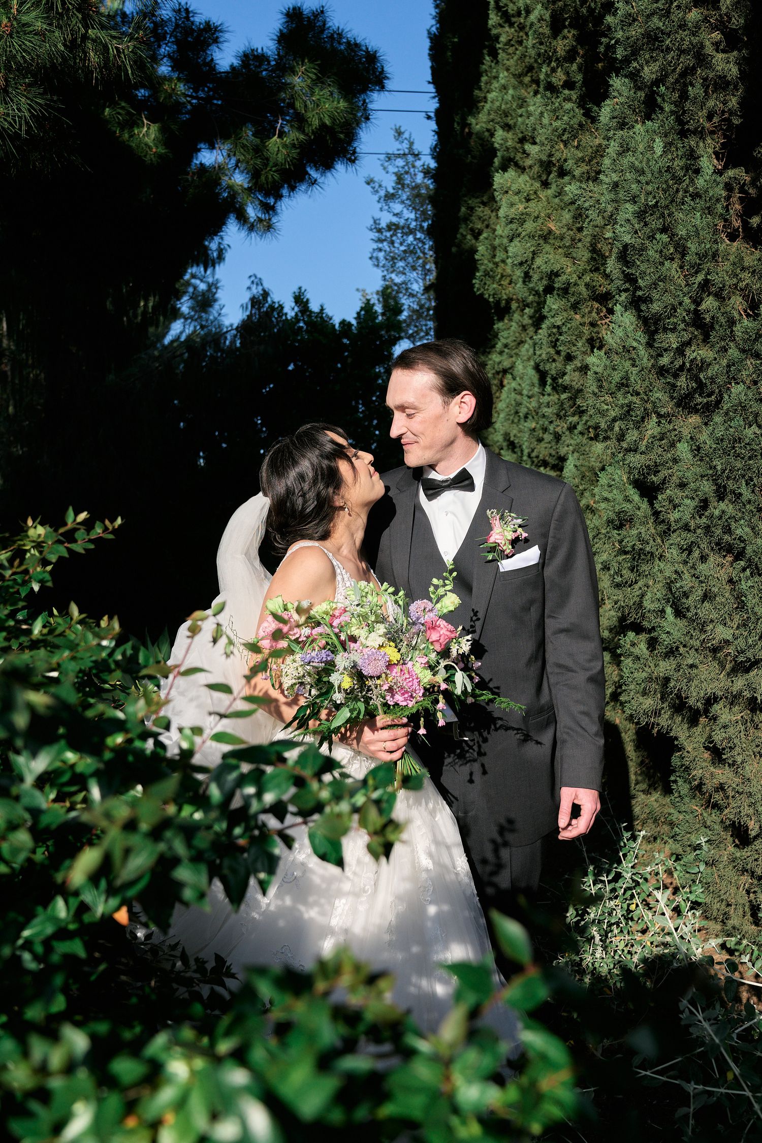 A romantic wedding portrait amongst cypress trees and garden foliage with floral bouquet.