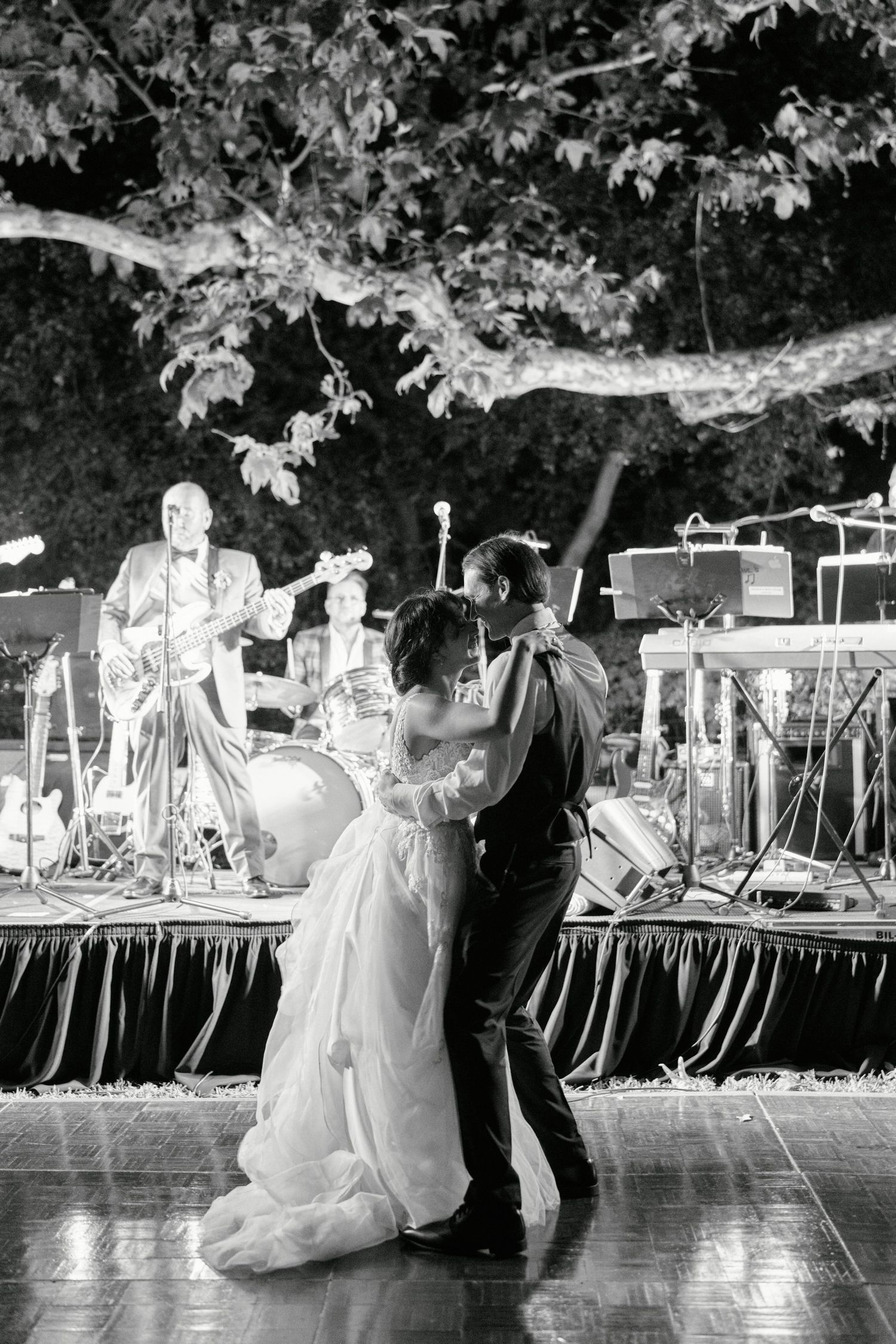 A couple shares their first dance at an outdoor wedding reception with a live band on stage.