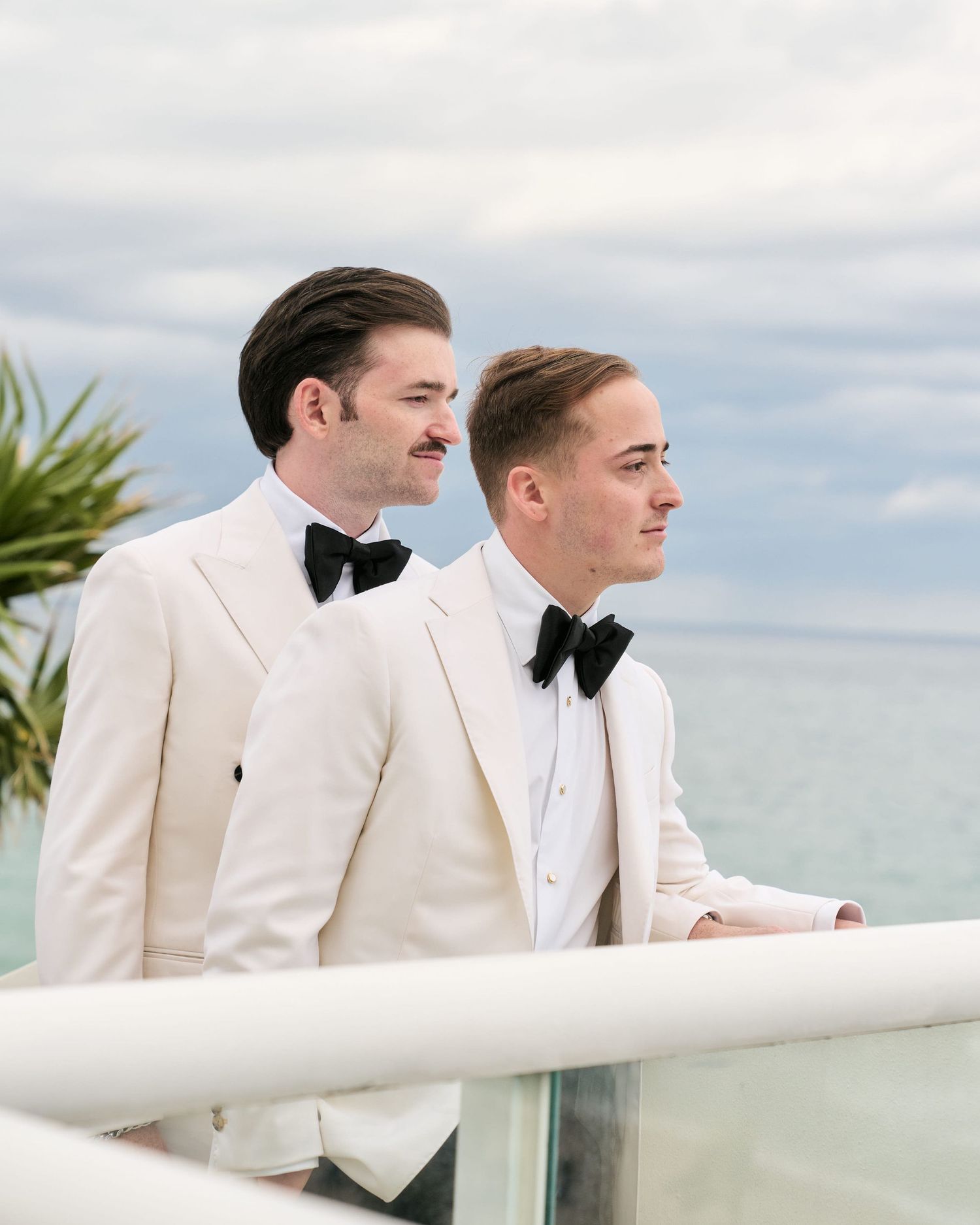 Two people in white tuxedos with black bow ties stand by an oceanfront railing.