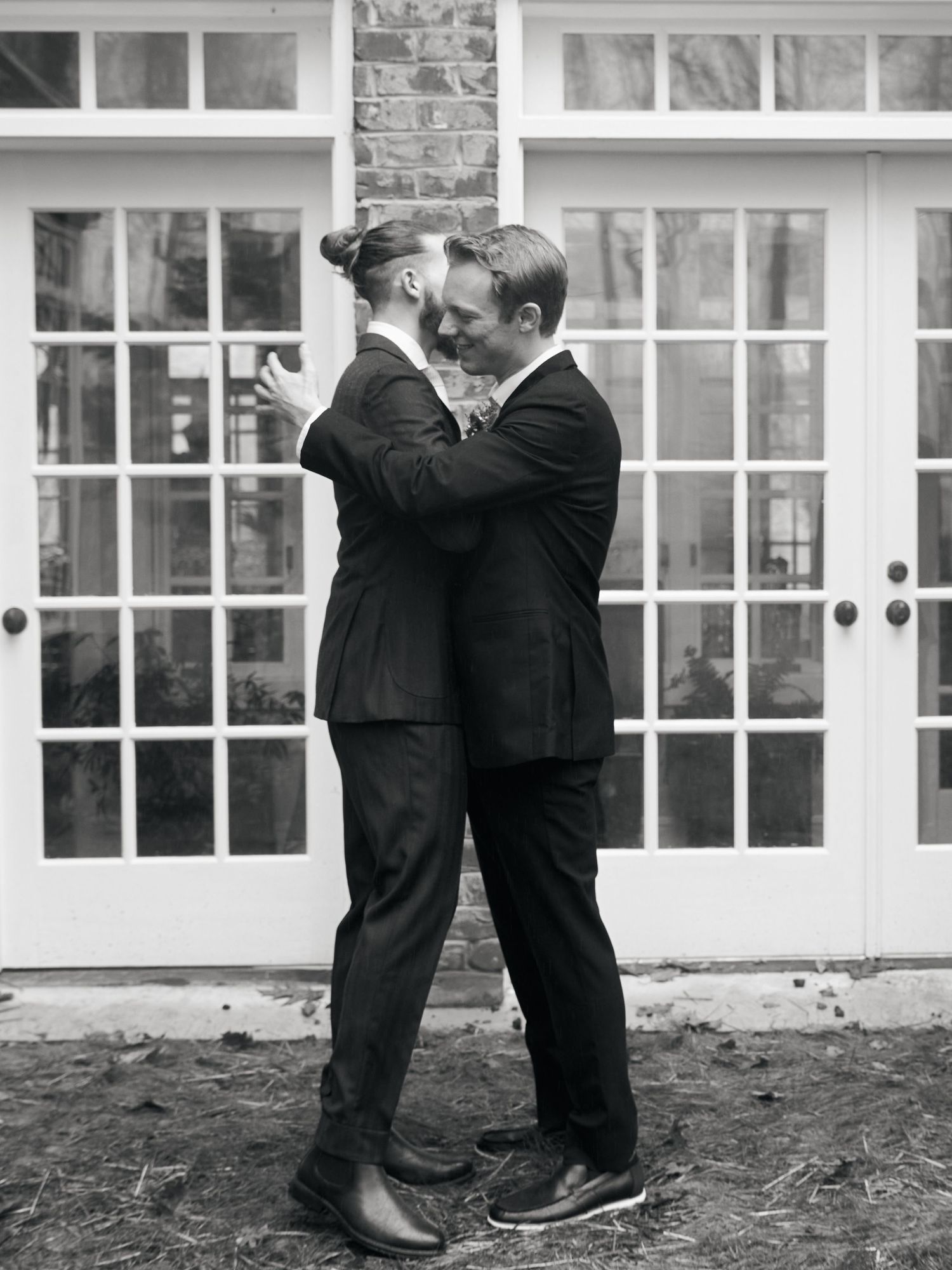 Black and white photo of two people in suits embracing outdoors in front of French doors.