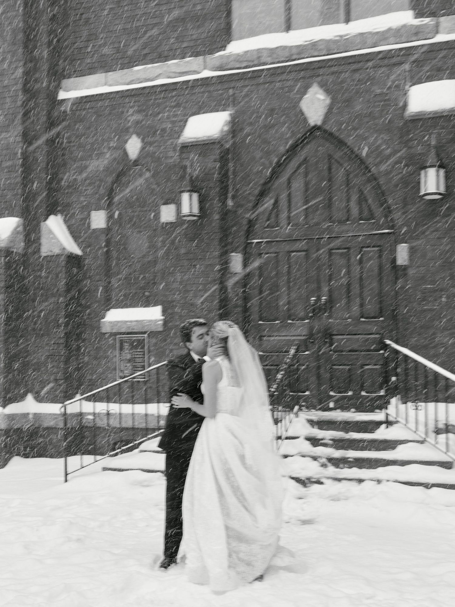 Black and white photo of bride and groom embracing in snowstorm outside historic church on winter wedding day.