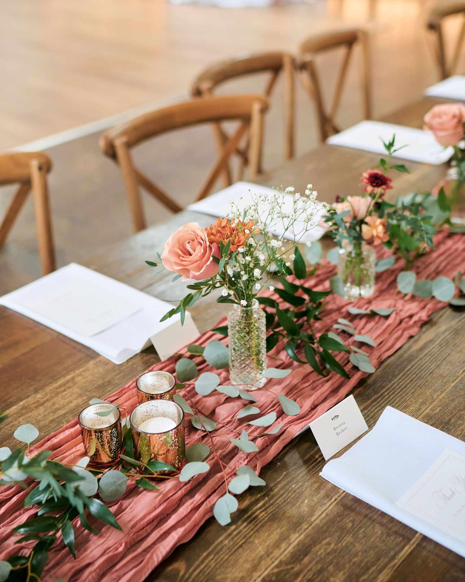 Rustic wedding table with dusty rose cheesecloth runner, eucalyptus greenery, pink roses, and gold candle holders on wood table.