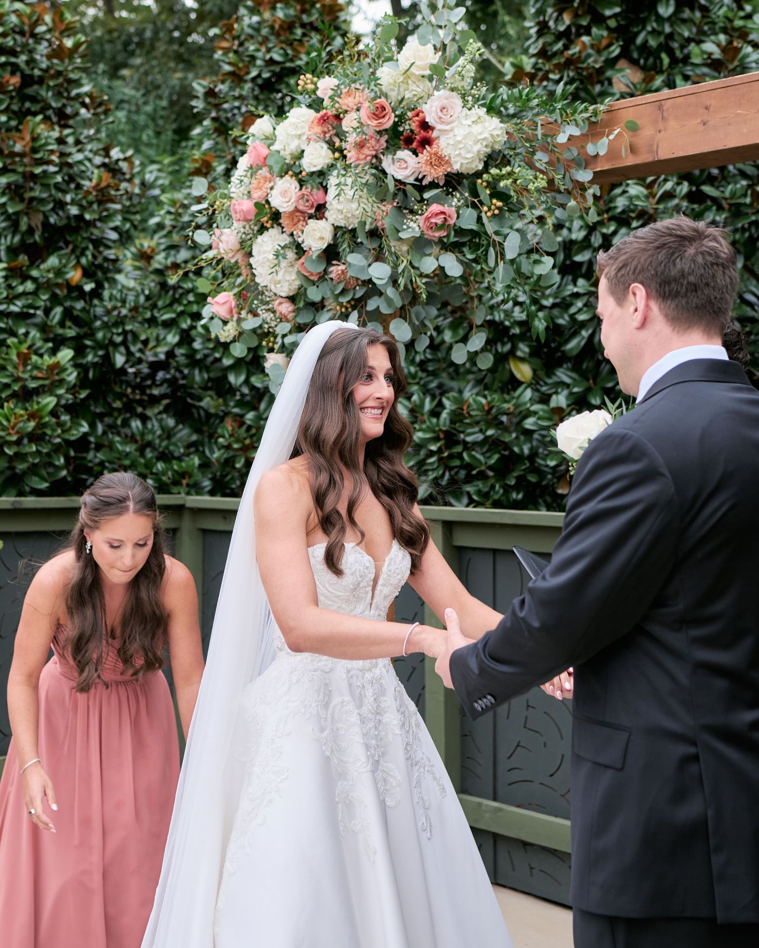Smiling bride in lace gown holds grooms hands at outdoor wedding ceremony beneath floral arch.