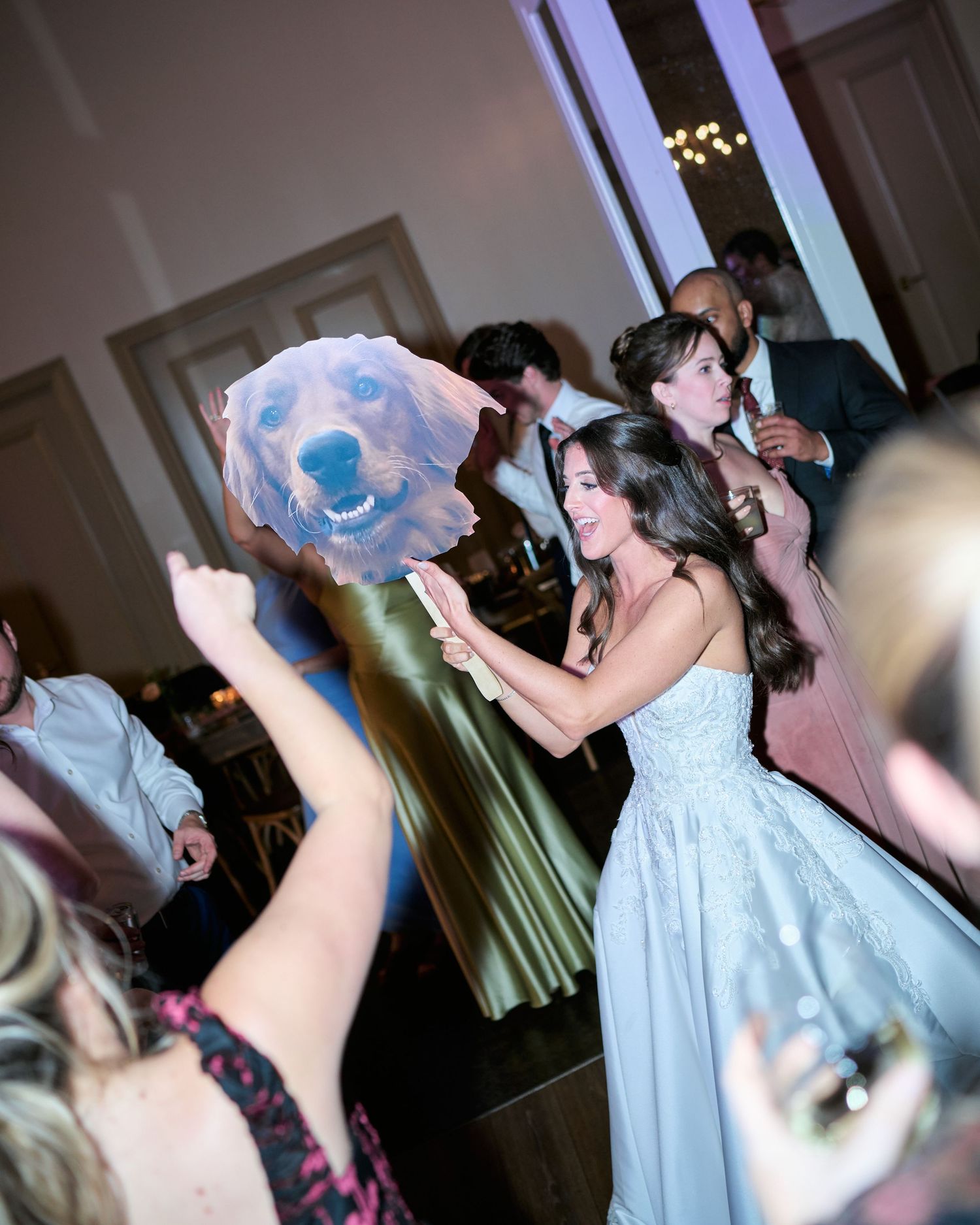 Bride in blue gown holds large dog portrait cutout while dancing with guests at elegant wedding reception.