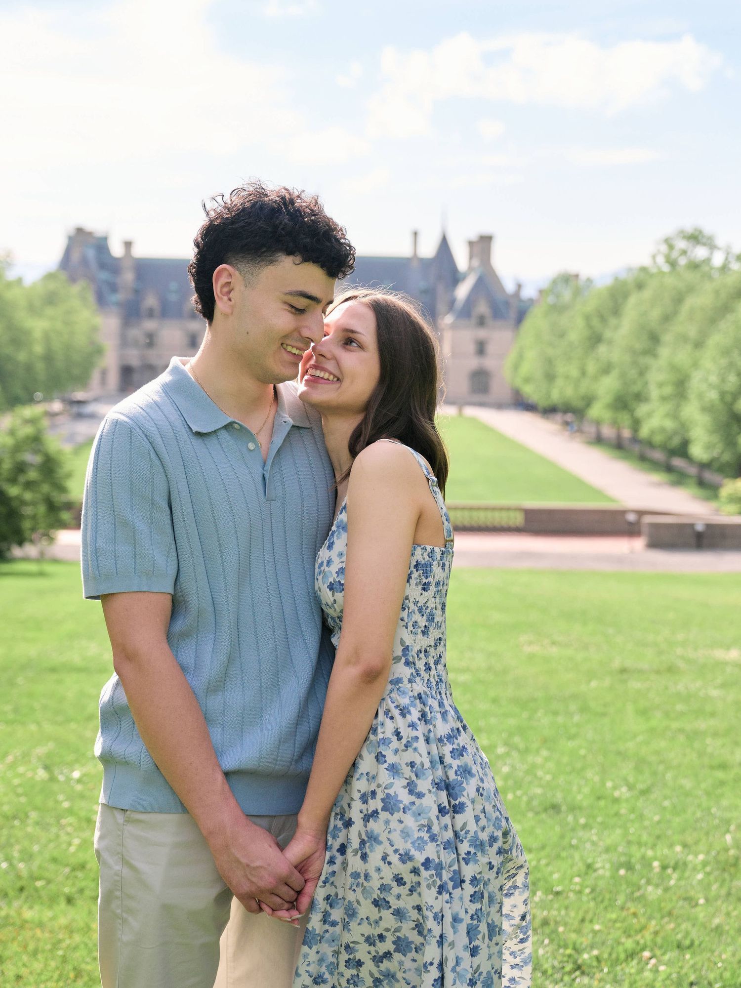 A young couple holds hands and smiles on a green lawn with a grand estate building in the background.