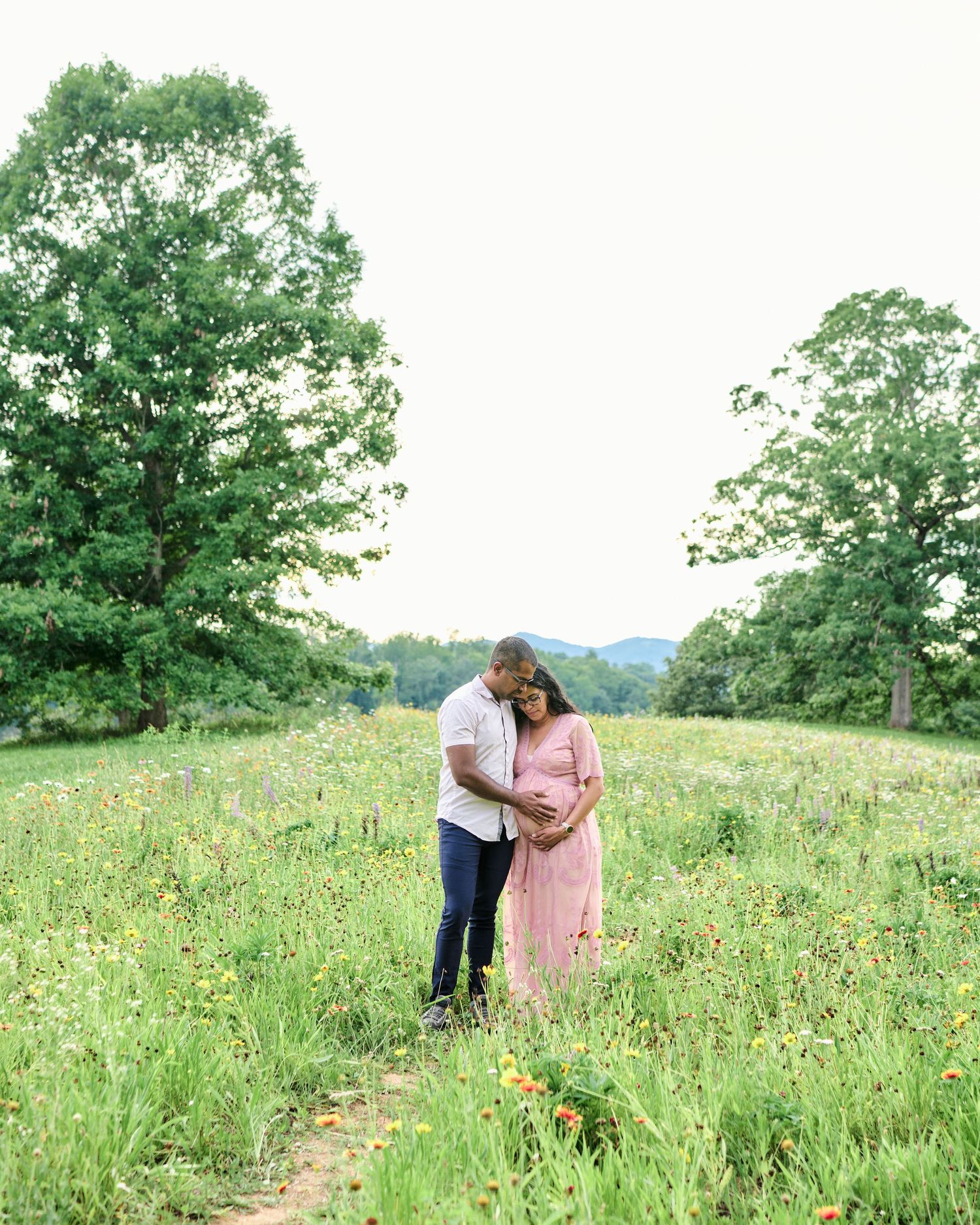 Couple embracing in a wildflower meadow during maternity photo session with lush green trees and mountains in background.