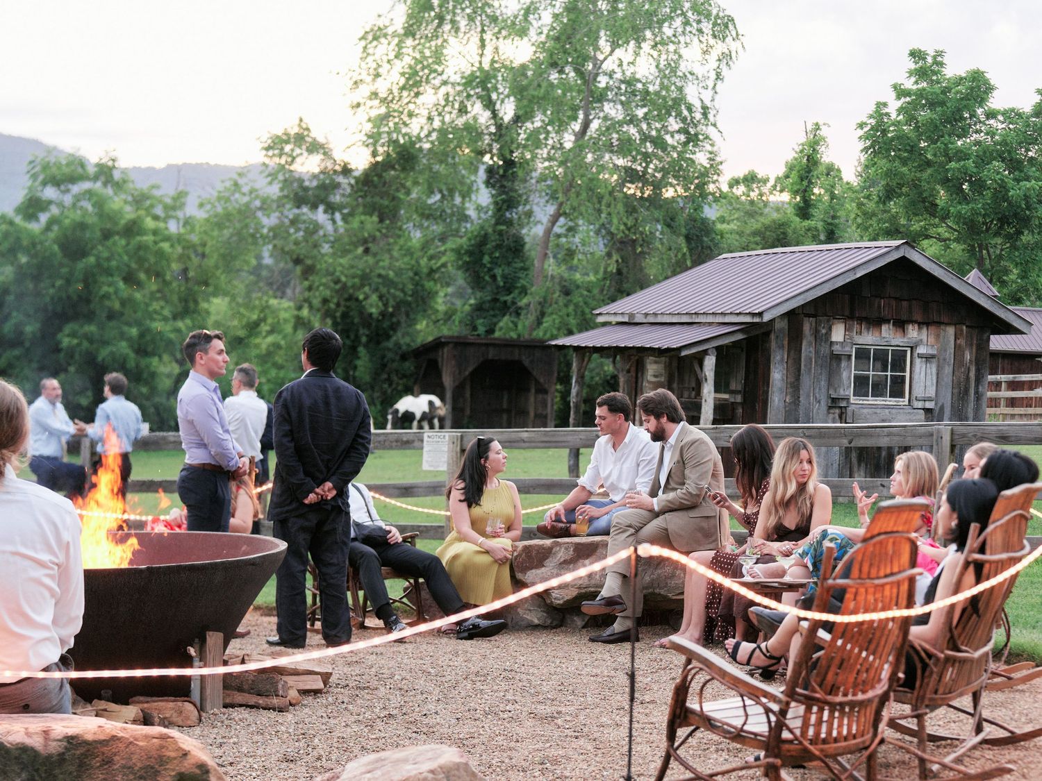 People gather around a rustic fire pit with chairs and a wooden cabin in the background at an outdoor event.