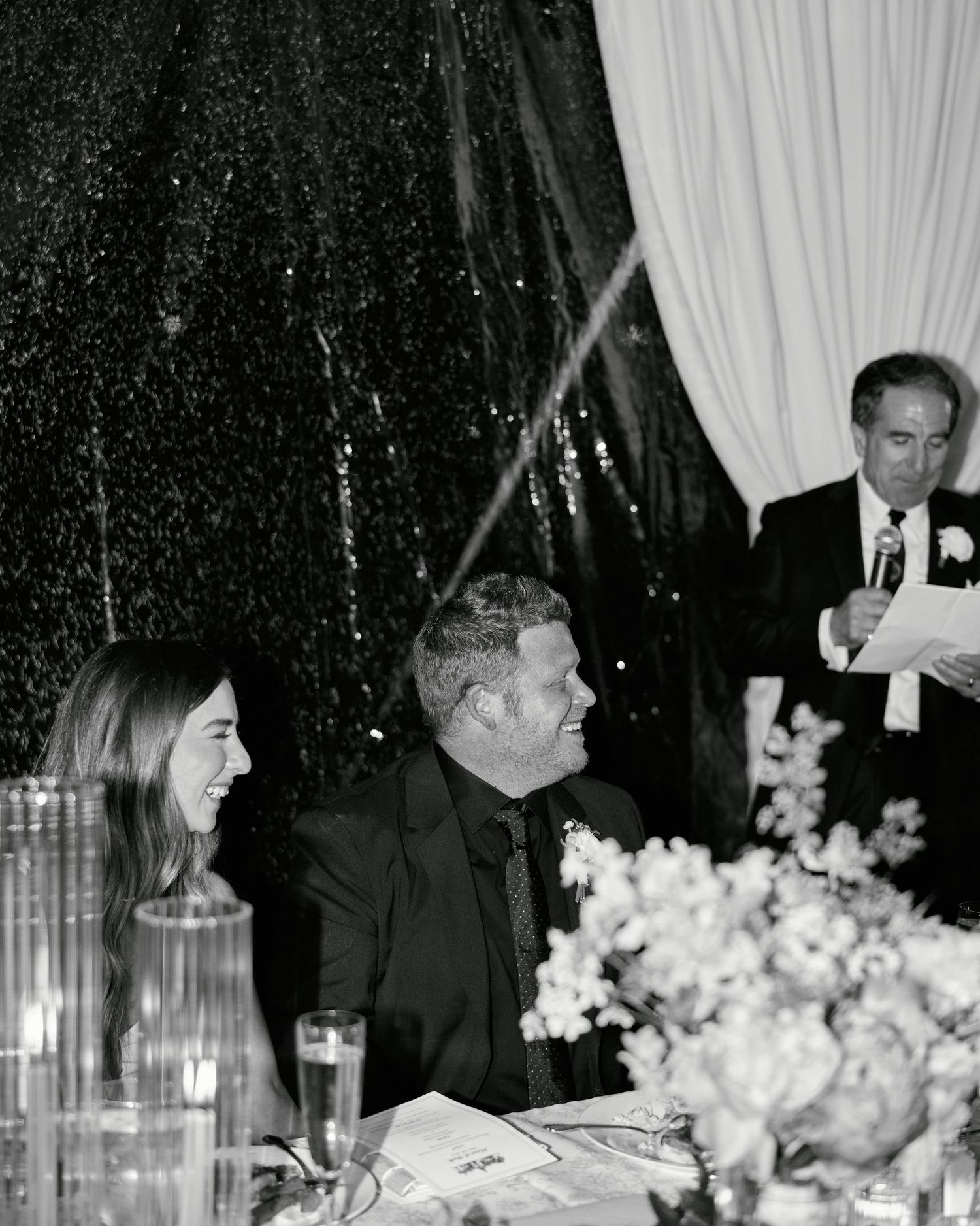 Black and white photo of guests at a formal event with floral centerpieces and white draping in the background.
