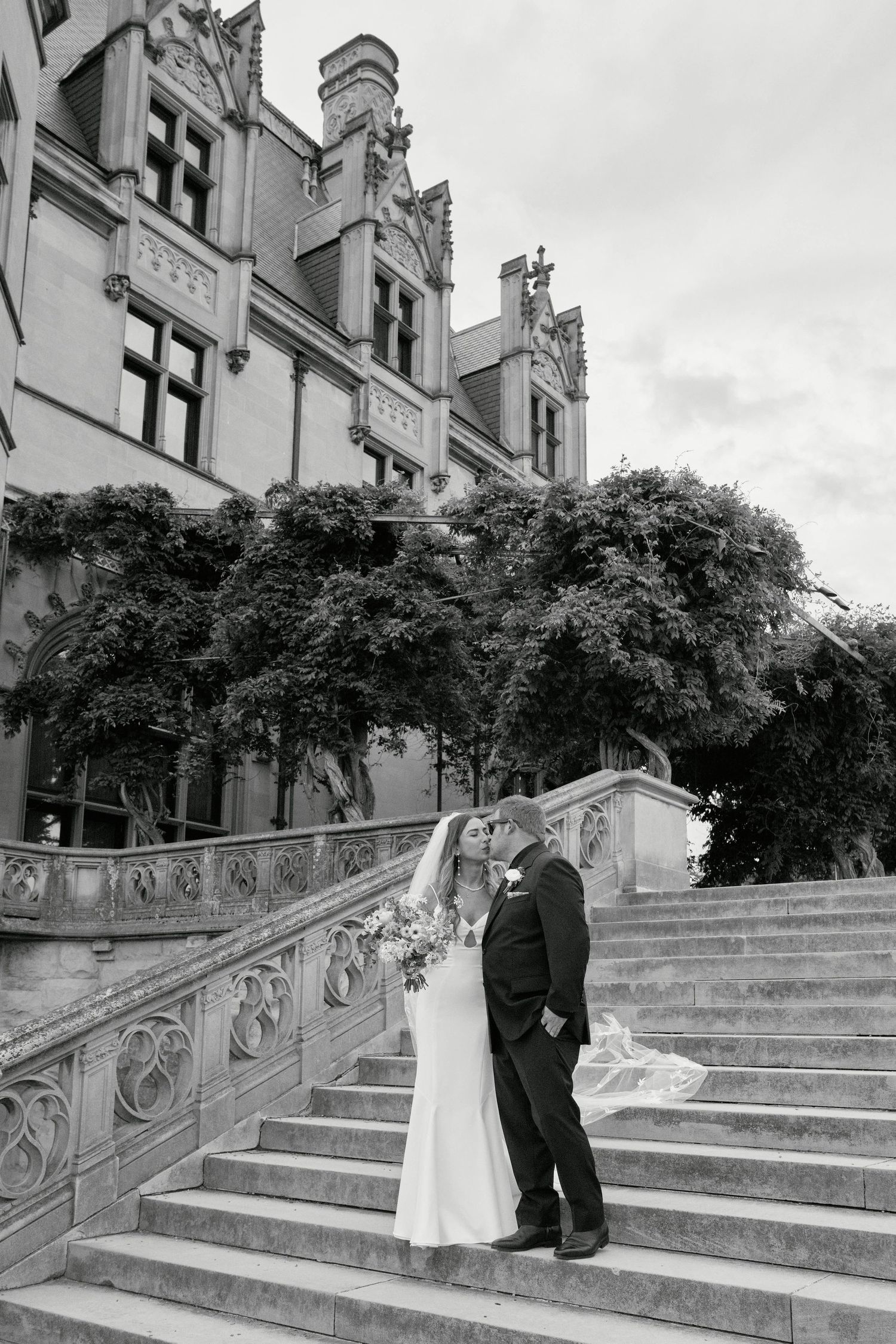A couple ascends an elegant stone staircase with ornate railings outside a historic building in black and white.