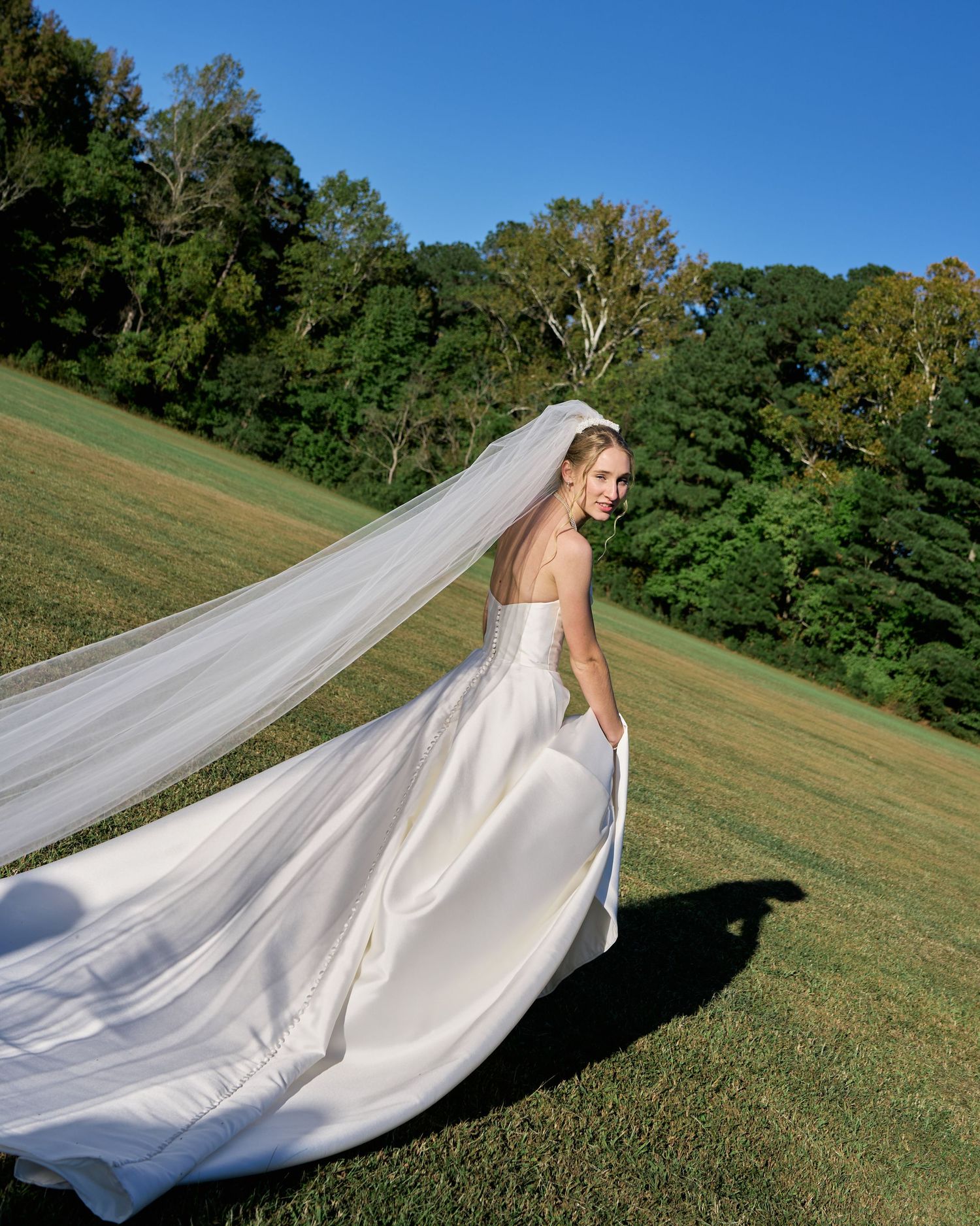 A flowing white wedding veil catches the wind as it trails behind someone on a grassy hillside.