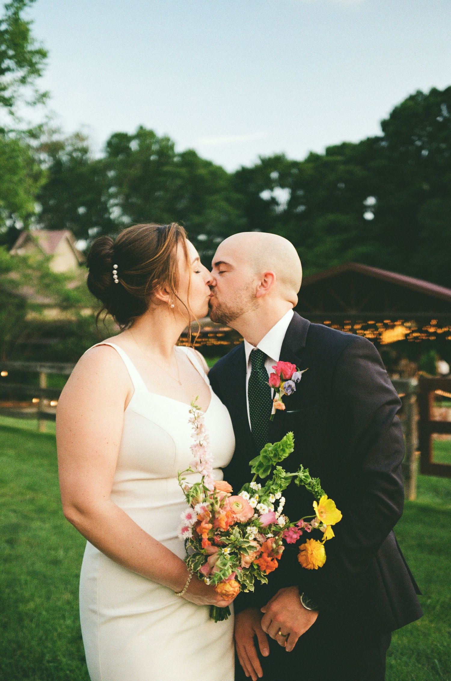 Newlywed couple sharing a kiss outdoors, bride holding colorful bouquet in white gown, groom in dark suit.