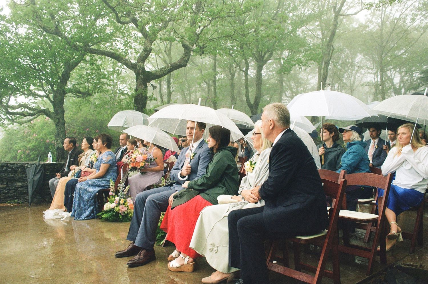 Guests holding white umbrellas sit on red chairs at an outdoor wedding ceremony on a rainy day.