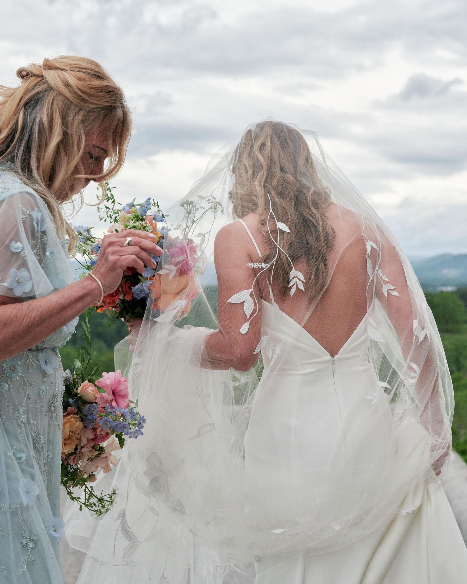 A bride in a white dress with a veil stands outdoors with someone in a light blue dress holding floral bouquets.